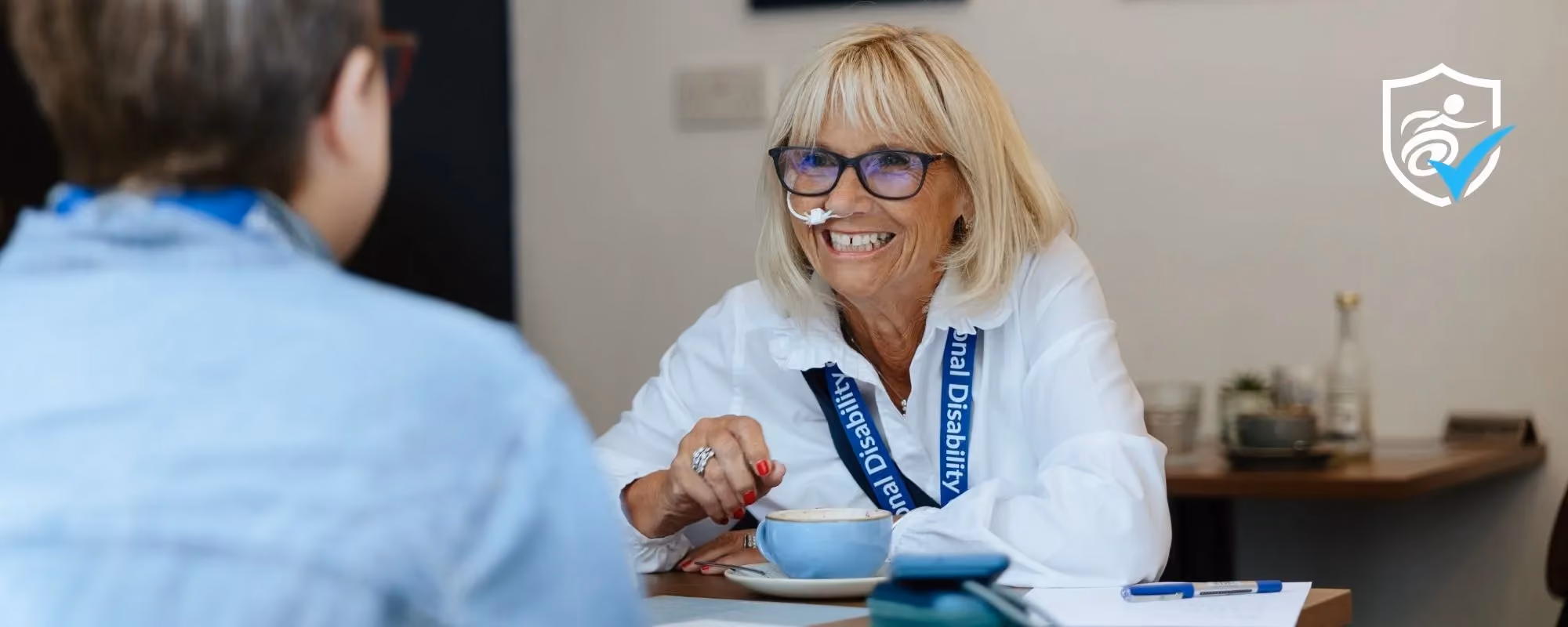 A woman wearing a National Disability Card lanyard sits in a cafe with a coffee