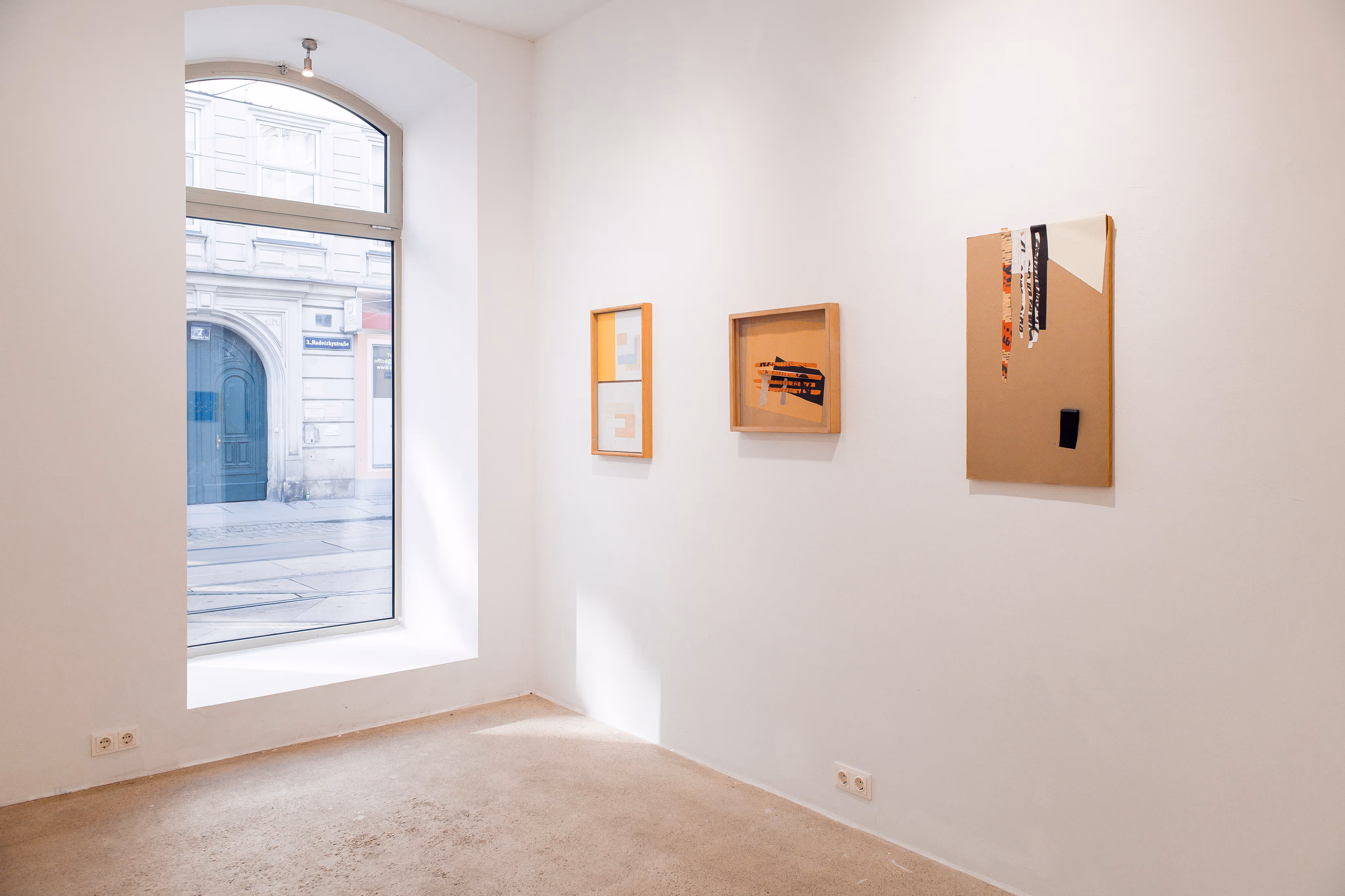 Exhibition view of Fritz Pechmann’s framed collage artworks in Vienna, 2025. Three warm-toned paper collages are displayed on a minimalist white wall: a vertical piece with layered paper squares, a horizontal collage with overlapping geometric fragments, and a tall cardboard collage with fabric and paper strips. Natural light from a large arched window highlights the textures and craftsmanship of these contemporary abstract collages.
