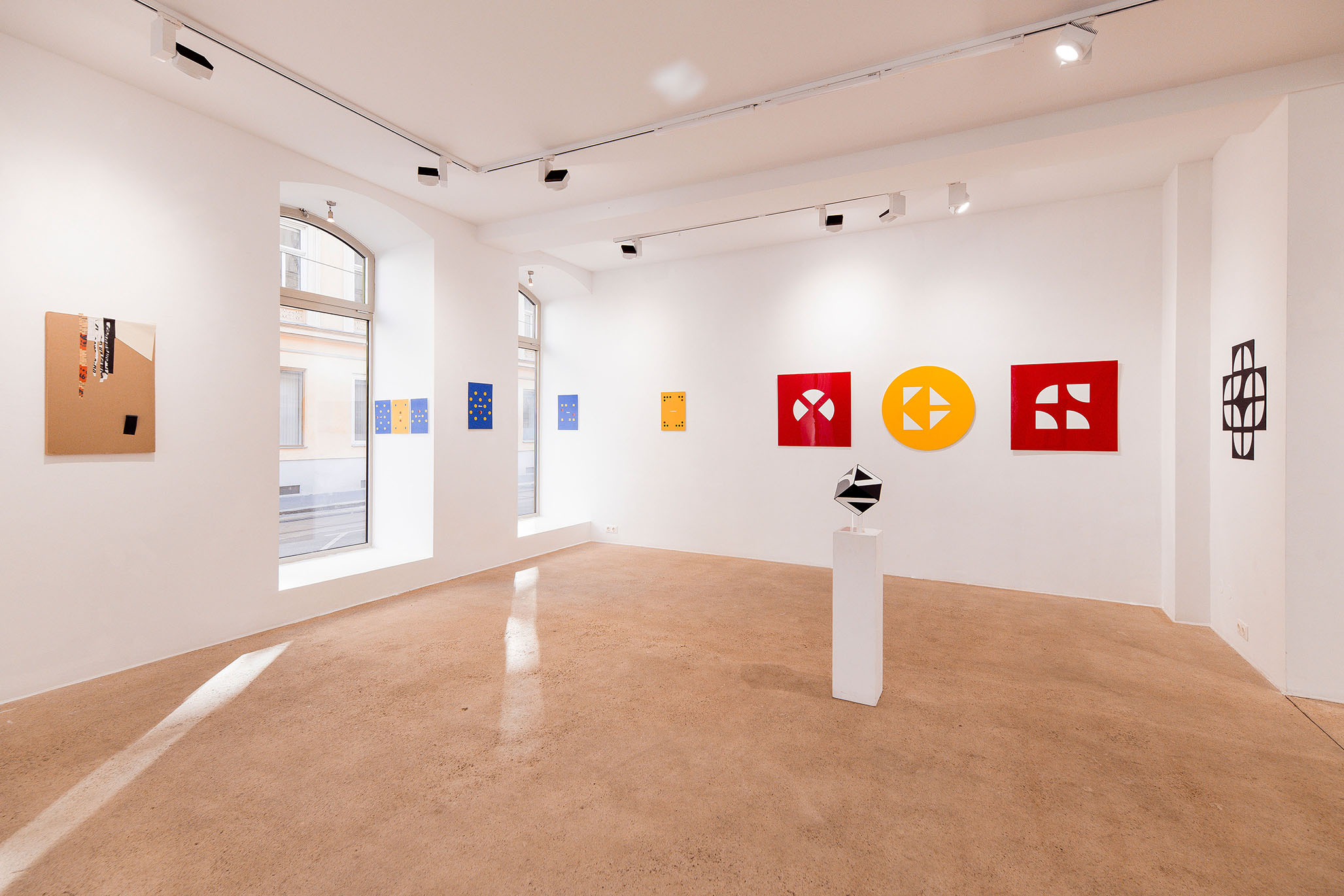 Gallery exhibition of Fritz Pechmann’s geometric concrete and abstract artworks, featuring colorful square and circular panels in red, yellow, and blue, with a black-and-white geometric sculpture on a white pedestal in a bright, minimal gallery space.