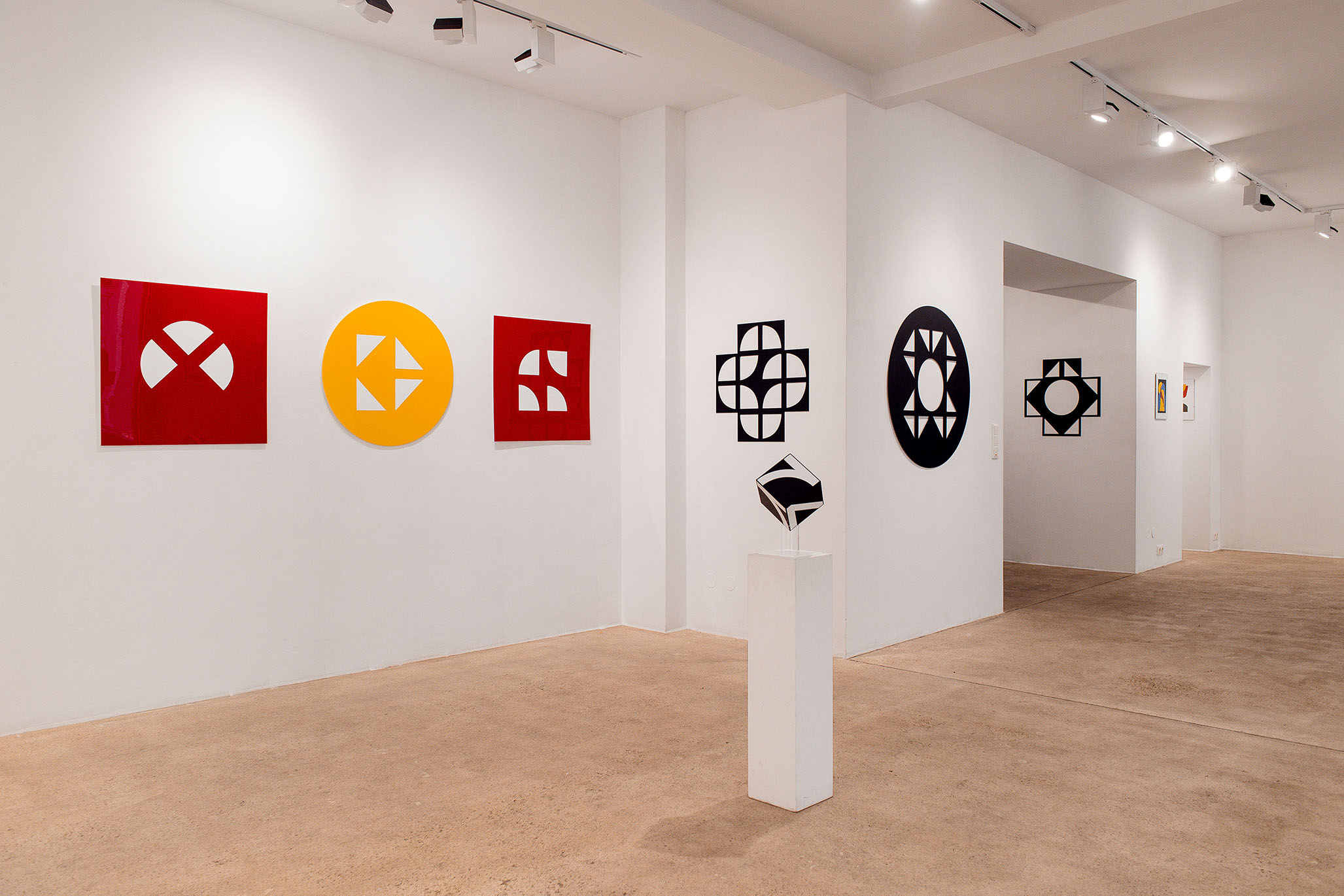 Exhibition of Fritz Pechmann’s geometric abstract artworks, featuring red and yellow panels with cut-out shapes, black circular and cross-like wall pieces, and a small black-and-white geometric sculpture on a white pedestal in a minimal gallery space.