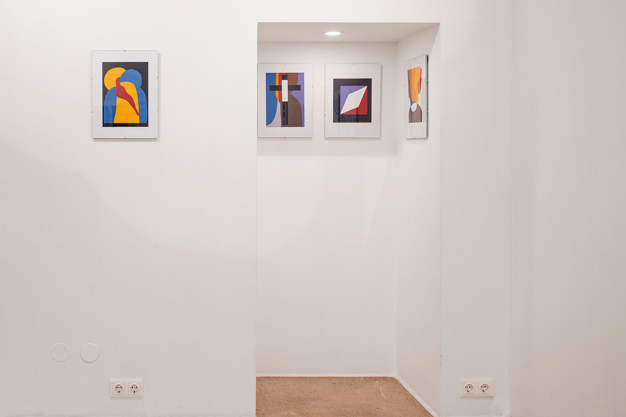 Minimalist gallery wall displaying Fritz Pechmann’s abstract Christian iconography, featuring small framed geometric artworks in blue, yellow, red, and black, arranged in a recessed white niche.