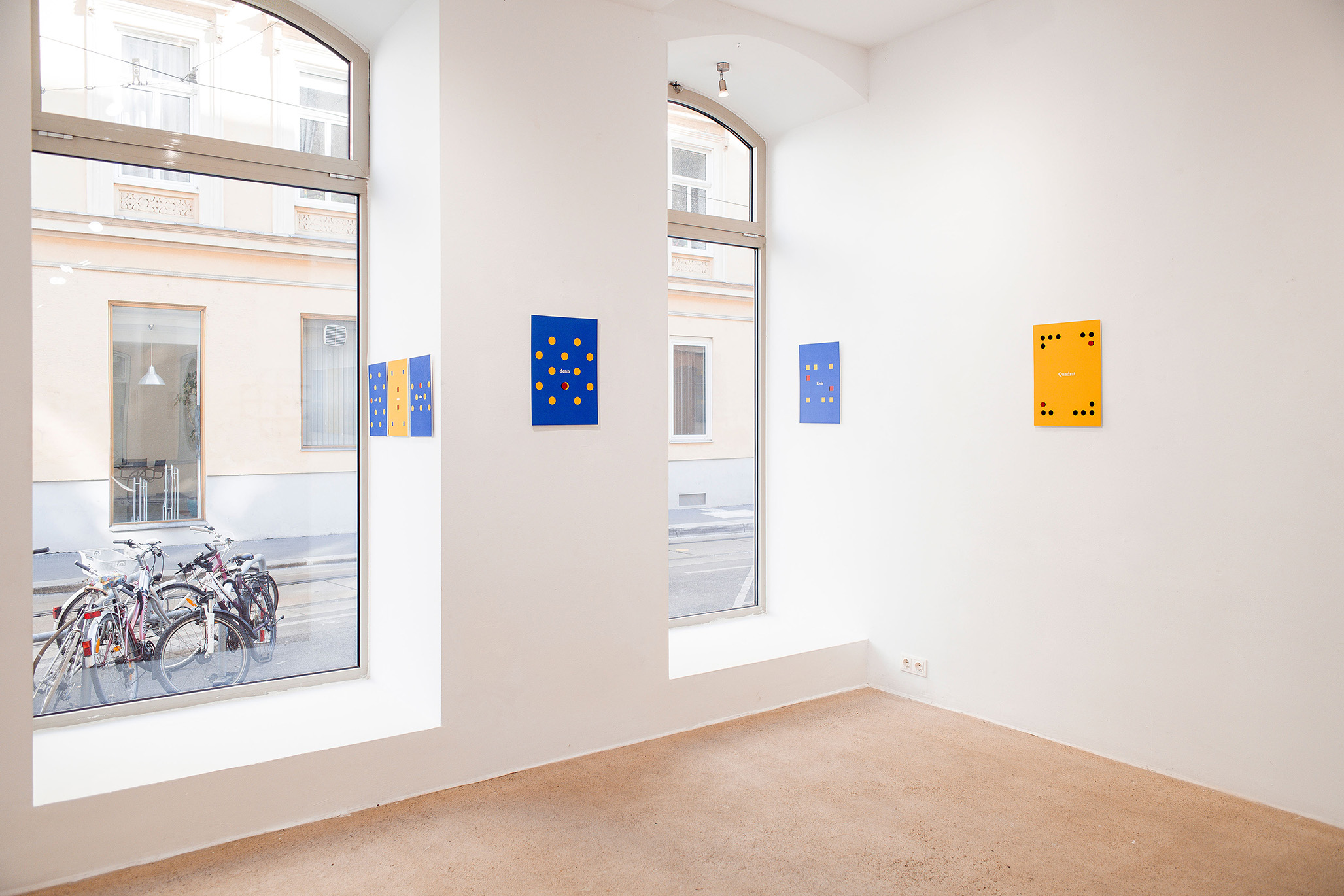 Exhibition view of Fritz Pechmann’s visual poetry series, featuring minimalist blue and yellow panels with dot compositions mounted on a white gallery wall, with large windows overlooking bicycles and a street outside.
