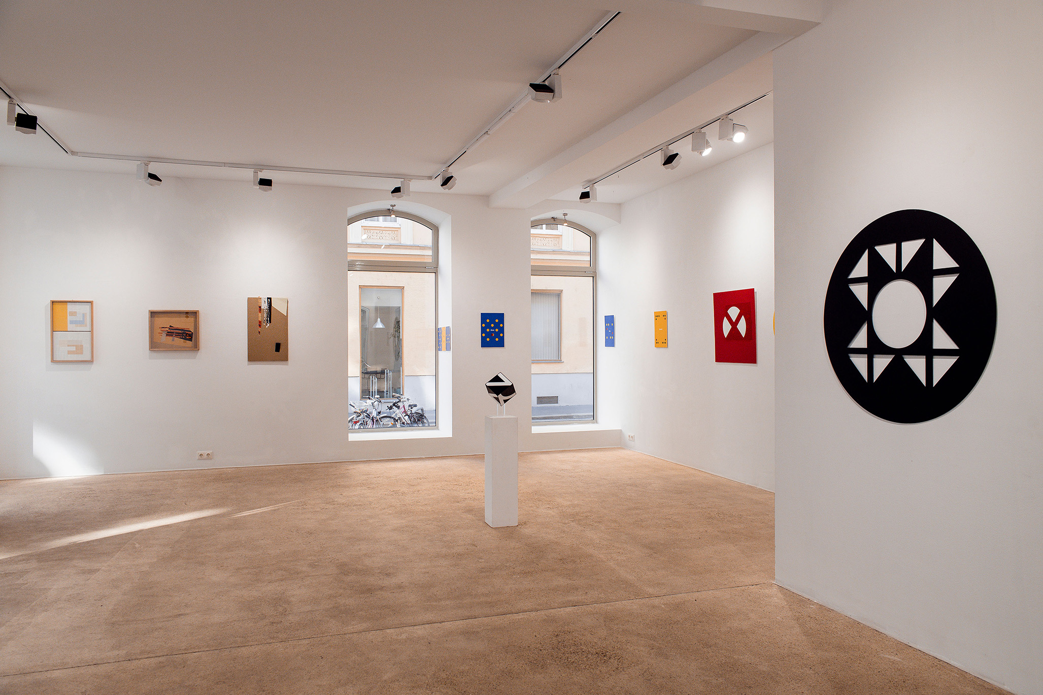 Wide view of a contemporary gallery exhibition featuring Fritz Pechmann’s geometric abstract artworks. A black circular wall piece with triangular cut-outs dominates the right, while colorful minimalist panels in red, yellow, and blue line the far wall. A black-and-white geometric sculpture stands on a white pedestal in the center, with natural light entering through large windows overlooking bicycles outside.