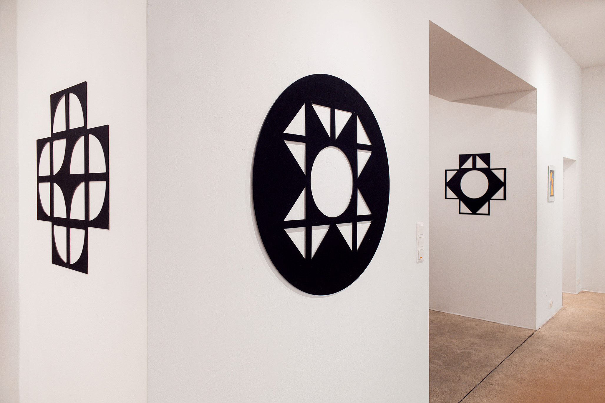 Gallery view of Fritz Pechmann’s black geometric wall objects: a cross-shaped piece on the left, an circular piece with triangular sunburst cut-outs in the center, and a square-cross design in the background.