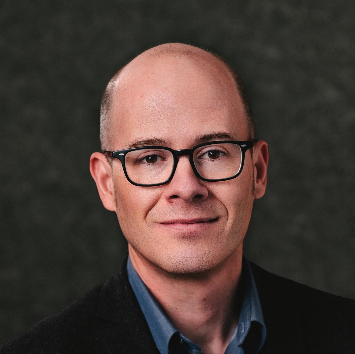 Headshot of a middle-aged man with a bald head wearing black rectangular glasses, a dark blazer, and a blue collared shirt against a dark blurred background.