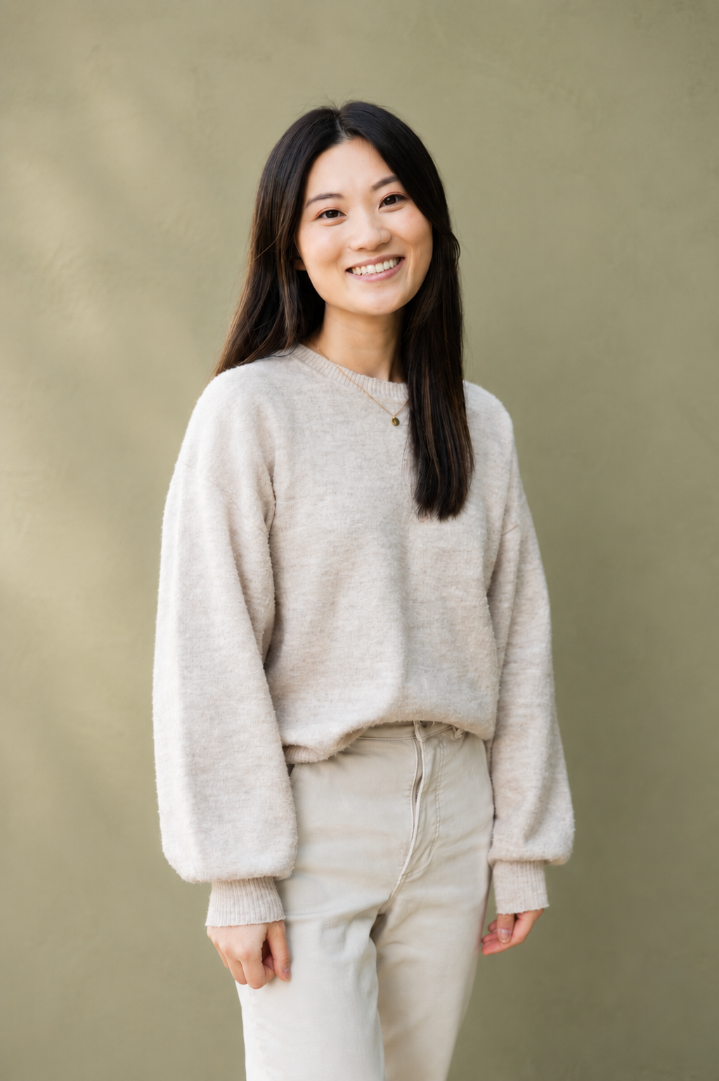 Smiling woman with long black hair wearing a beige sweater and pants against a plain background.