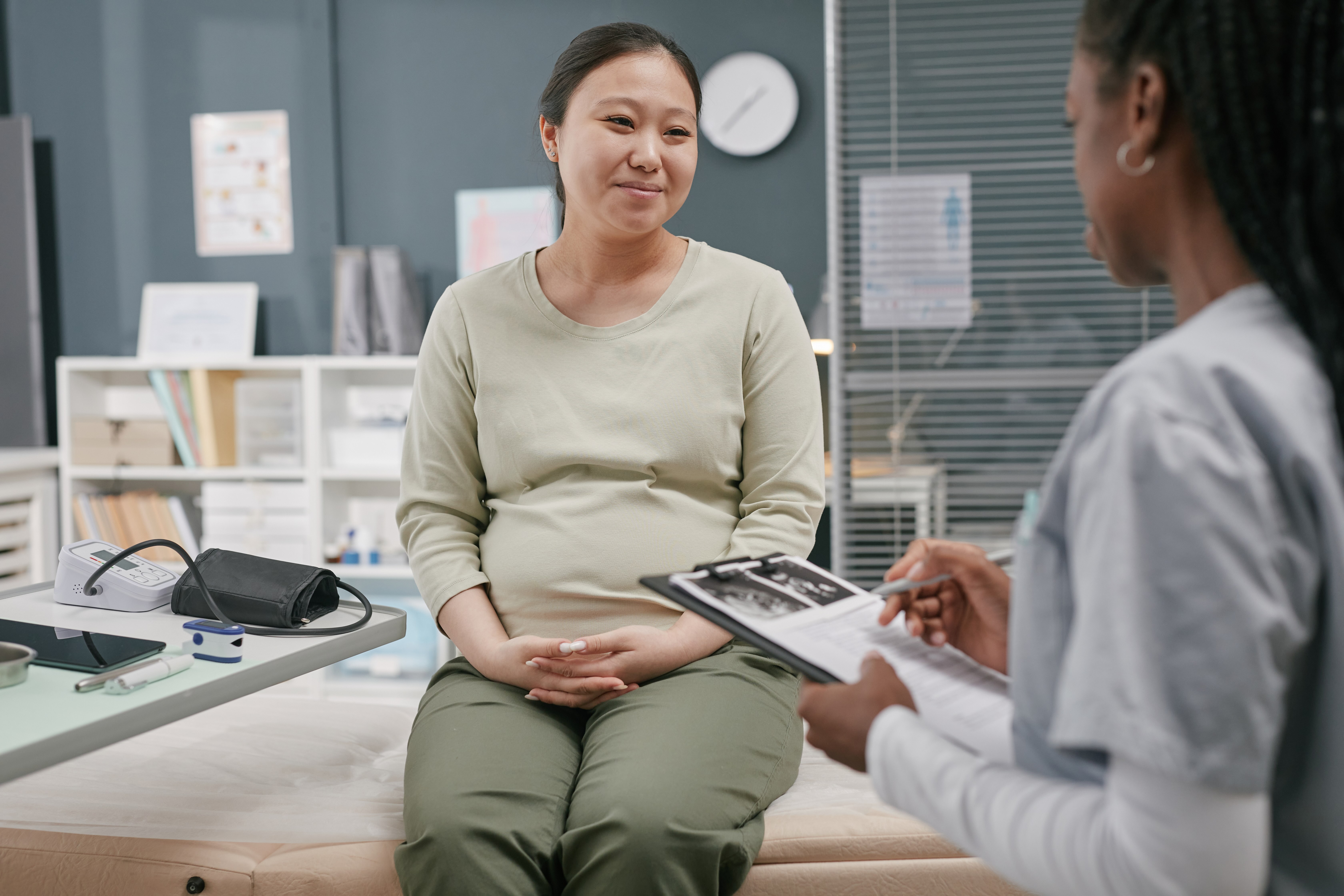 Pregnant woman sitting on a medical examination bed while a healthcare professional reviews ultrasound images and notes on a clipboard.