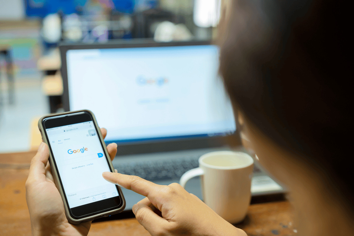 Person using Google search on a smartphone with a laptop open to Google search in the background.