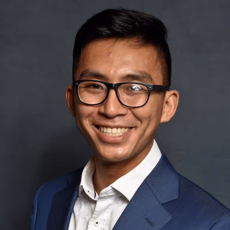 Smiling man with black glasses, wearing a white shirt and blue suit jacket against a dark background.