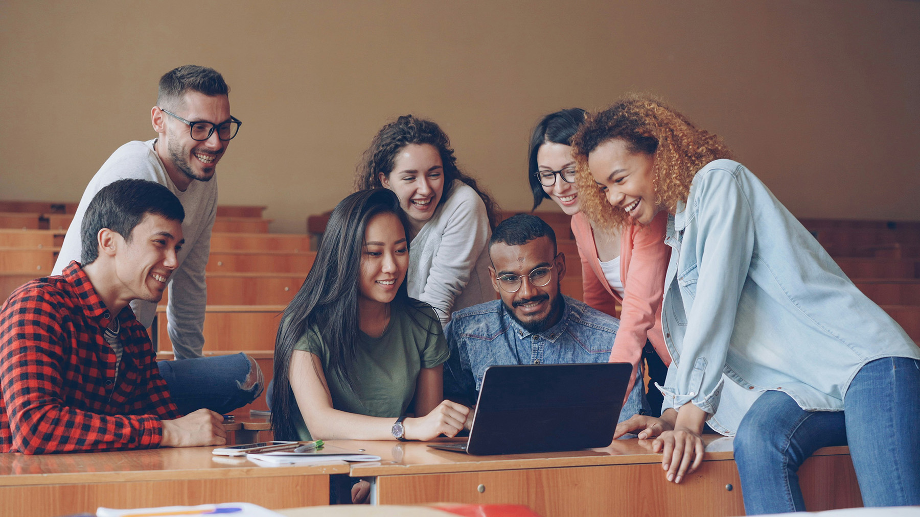 Group of diverse students smiling and looking at a laptop in a classroom.