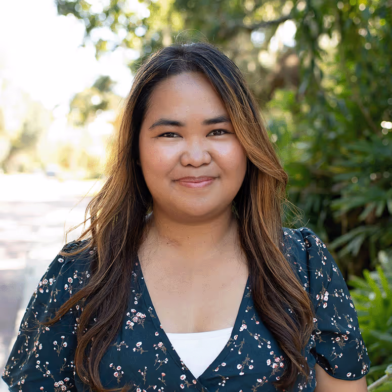 Woman with long brown hair wearing a dark floral dress standing outdoors in a sunlit green environment.