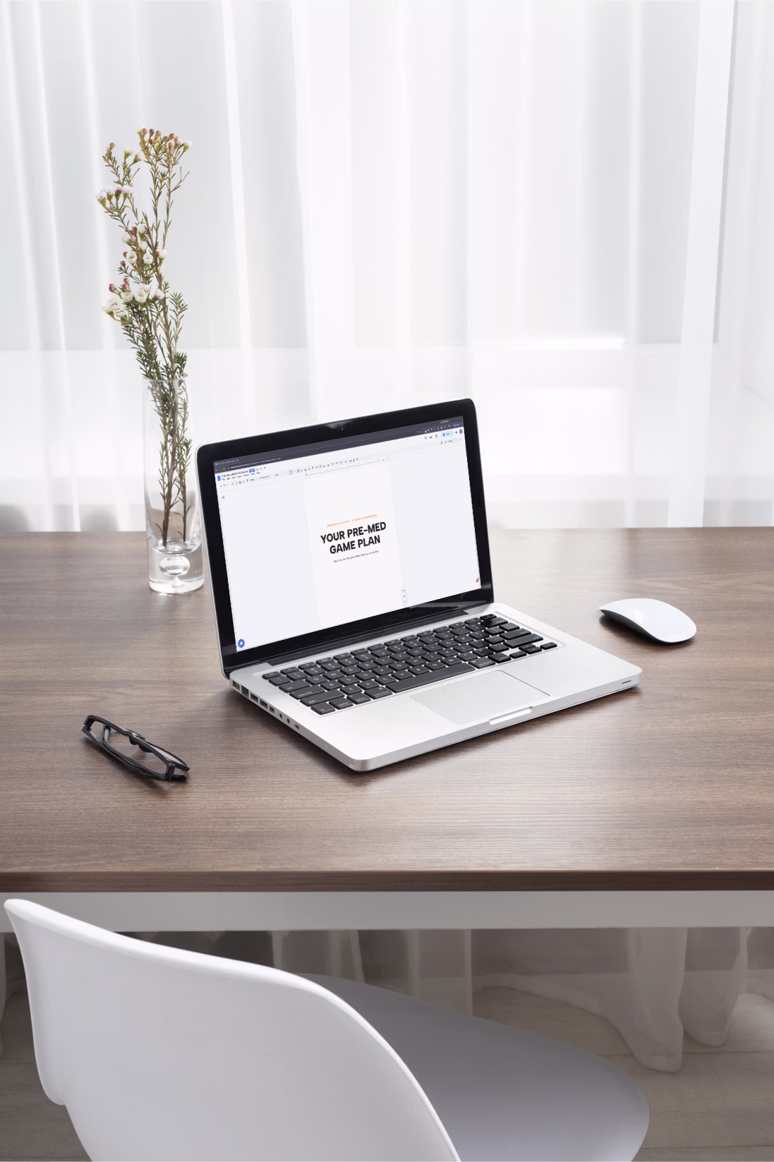 Laptop on a wooden desk displaying a document titled 'YOUR PRE-MED GAME PLAN' with a white mouse, glasses, and a vase with flowers nearby.