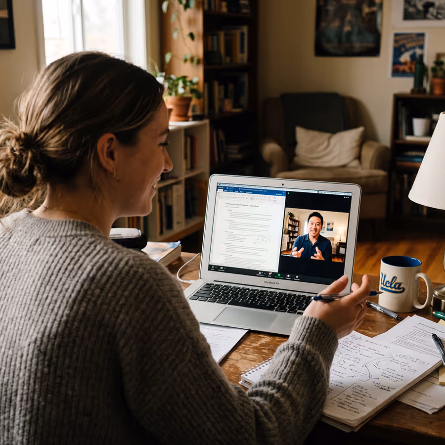 Woman in a gray sweater sits at a wooden desk engaged in a video call on a laptop with a man, surrounded by notebooks, papers, and a UCLA coffee mug.