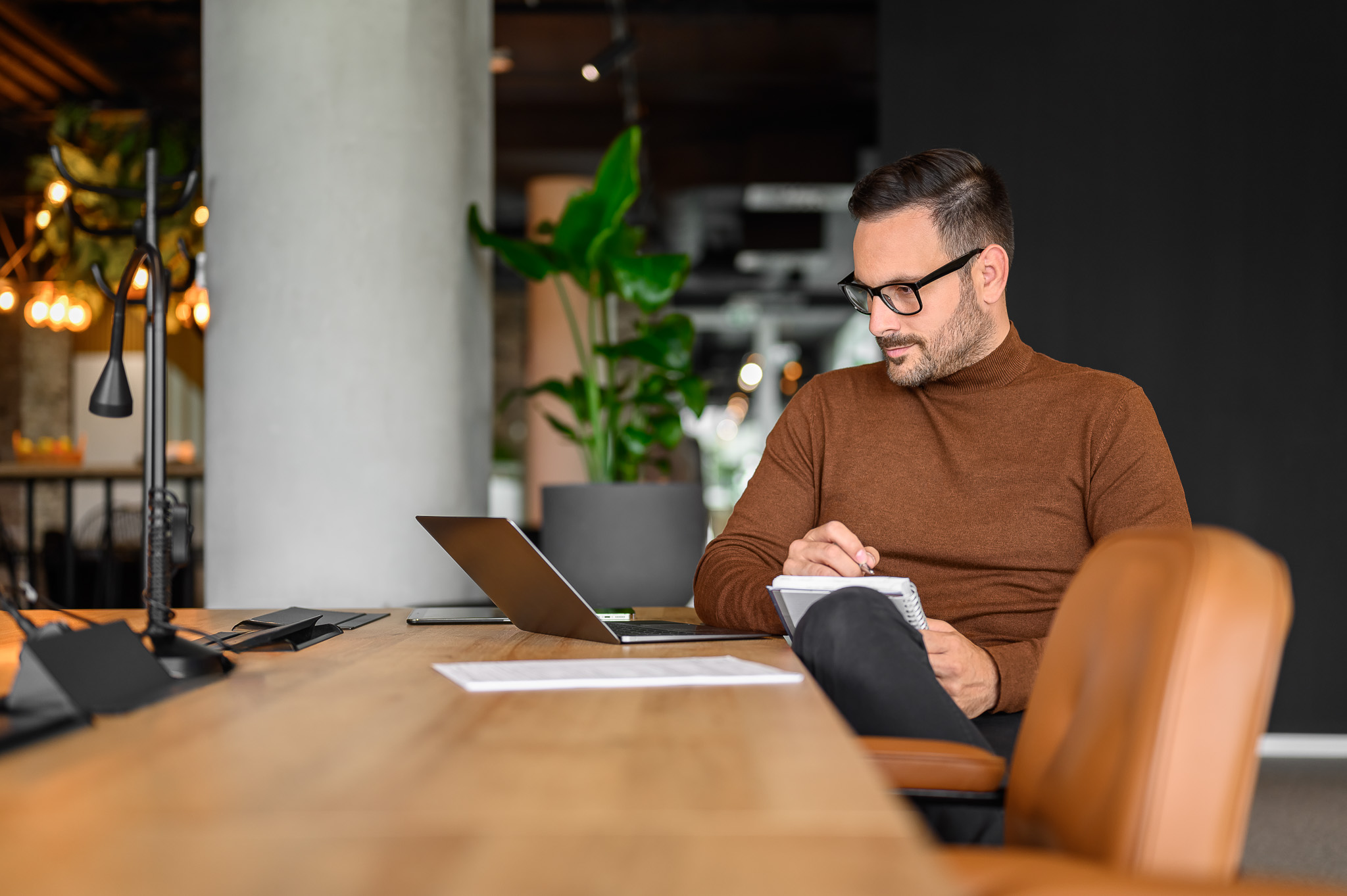 Man in a brown sweater taking notes at a modern office desk with a laptop.