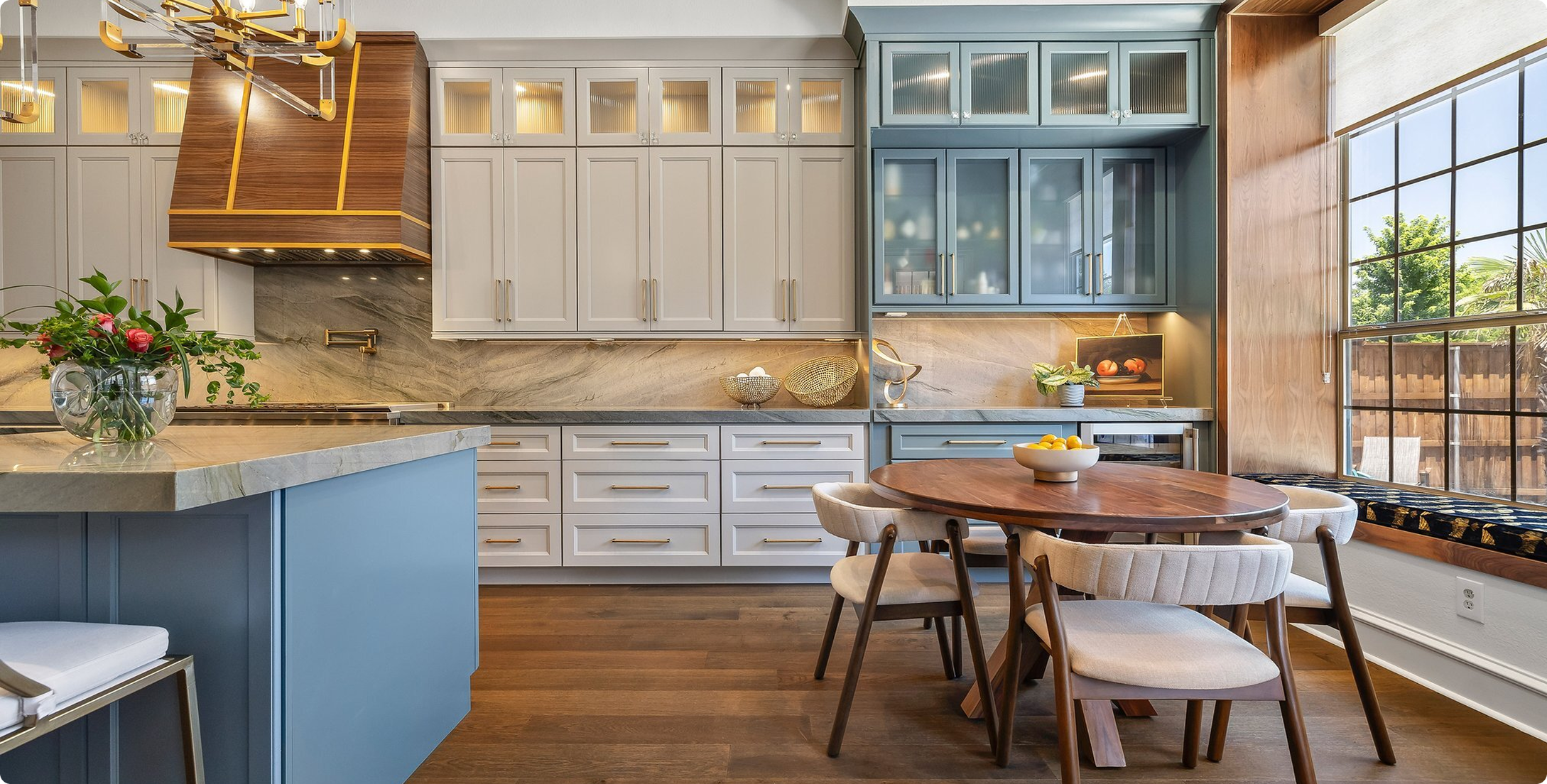 Modern kitchen with blue and white cabinetry, wooden round dining table with cream chairs, large window, and marble countertops.