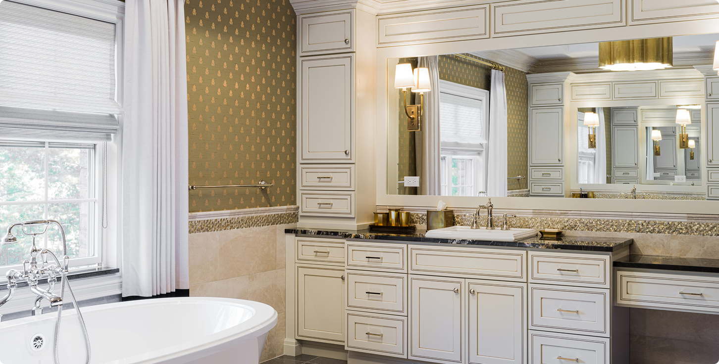 Elegant bathroom with a white freestanding bathtub, large mirror, beige patterned wallpaper, and cream cabinetry with black countertop.