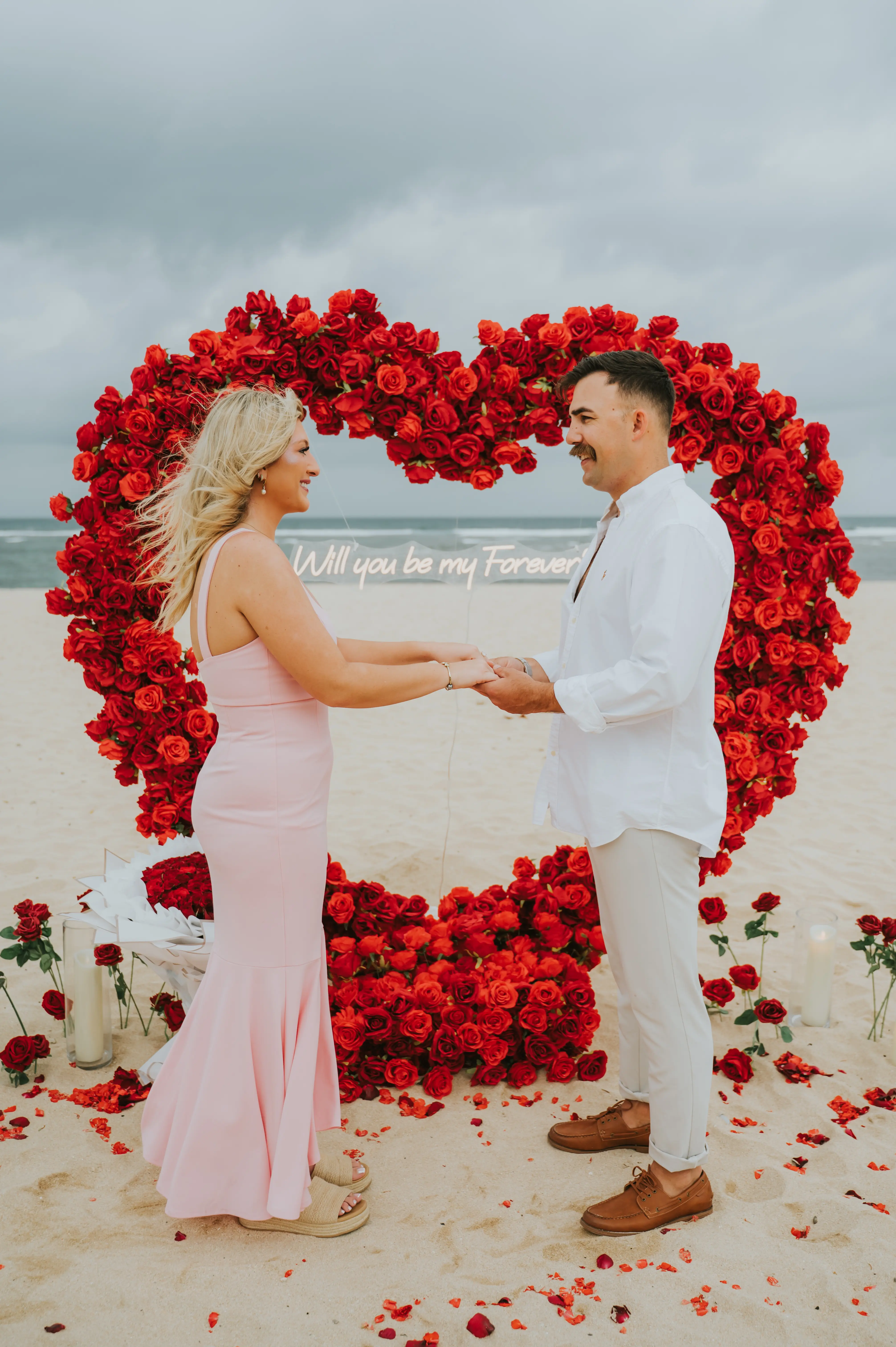 Engaged couple in front of red rose heart arch on the beach in Bali