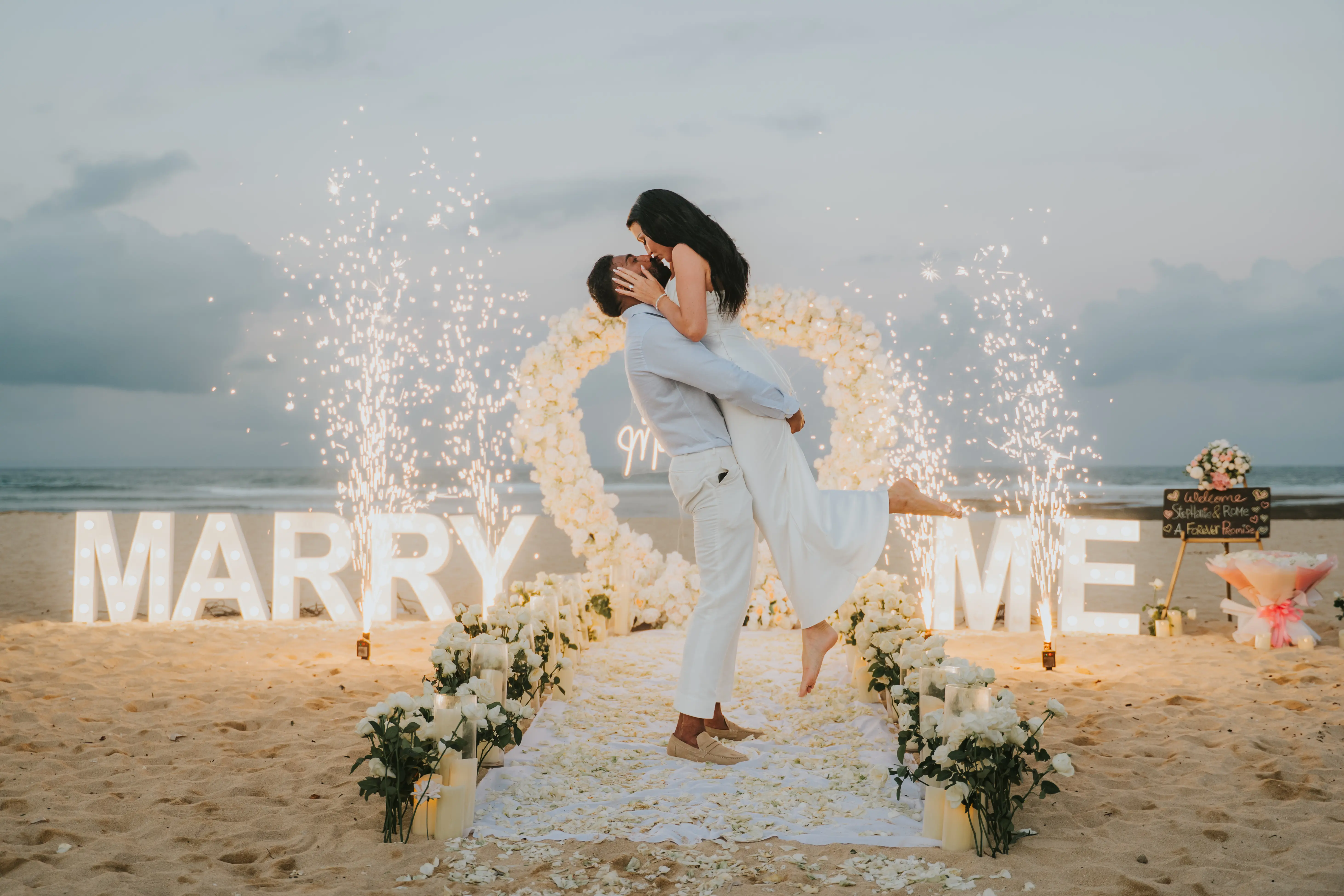 Groom lifting bride in front of “Marry Me” letters on the beach.
