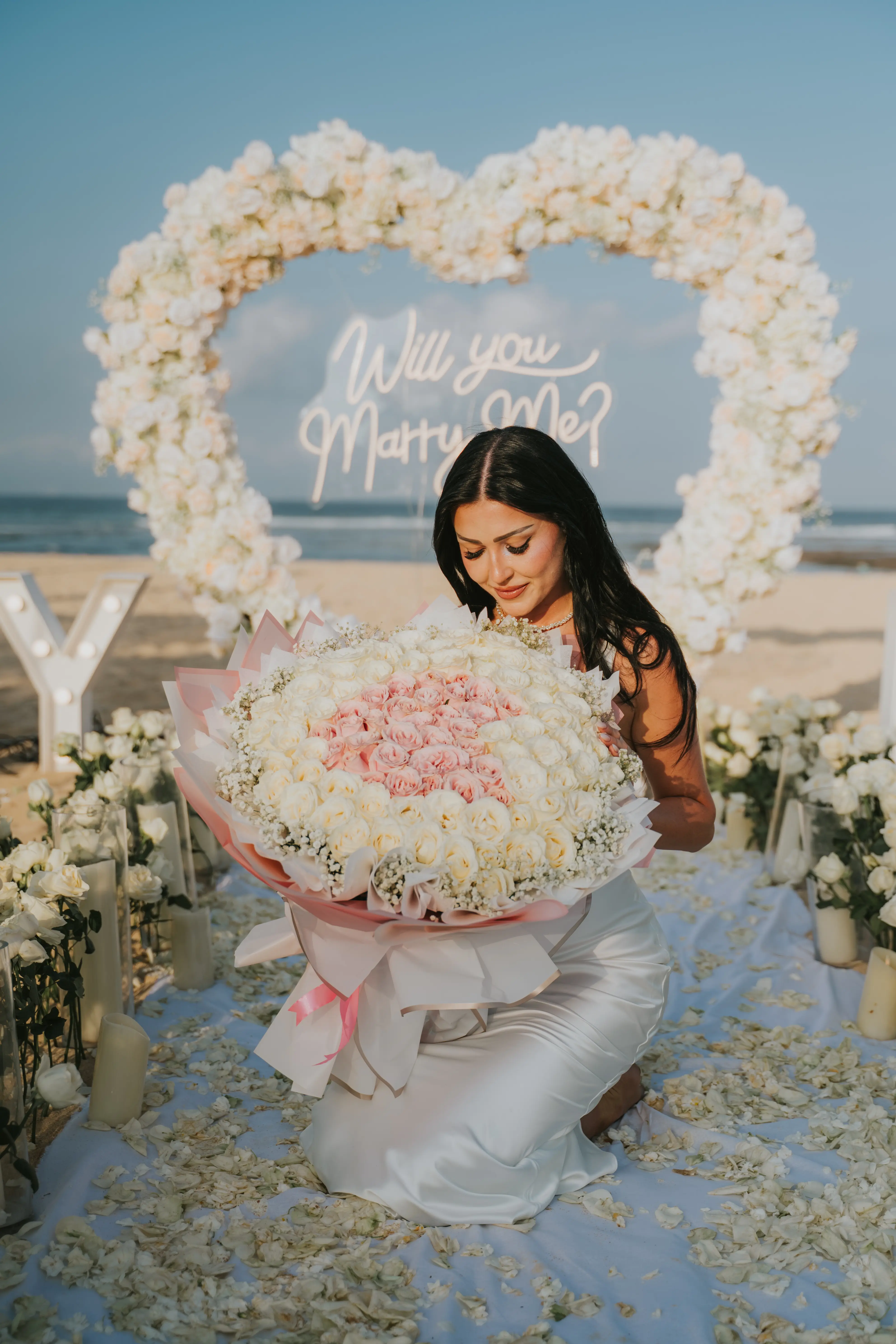 Bride sitting in front of floral arch holding bouquet of white roses.