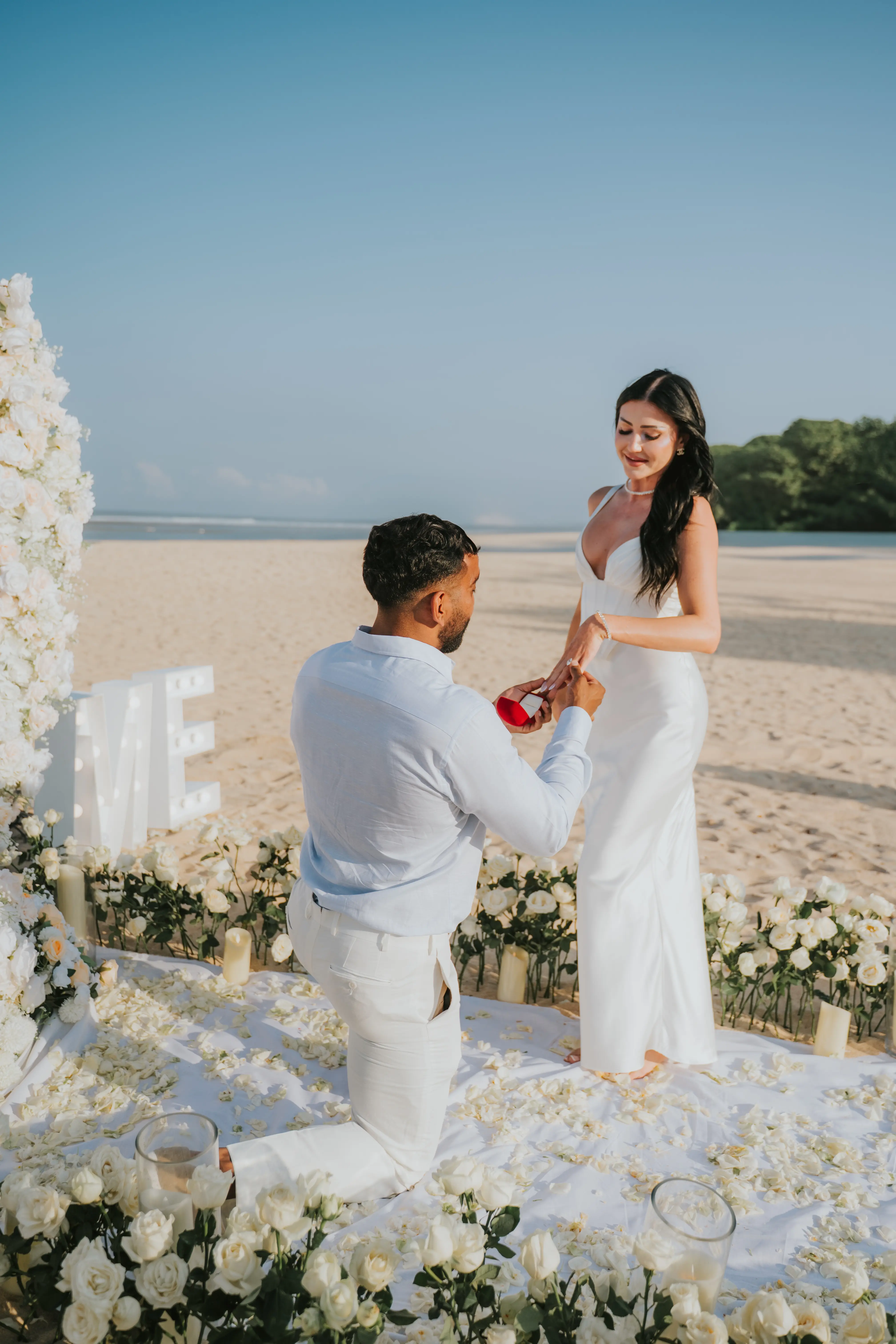 Groom proposing to bride on white-themed beach setup.