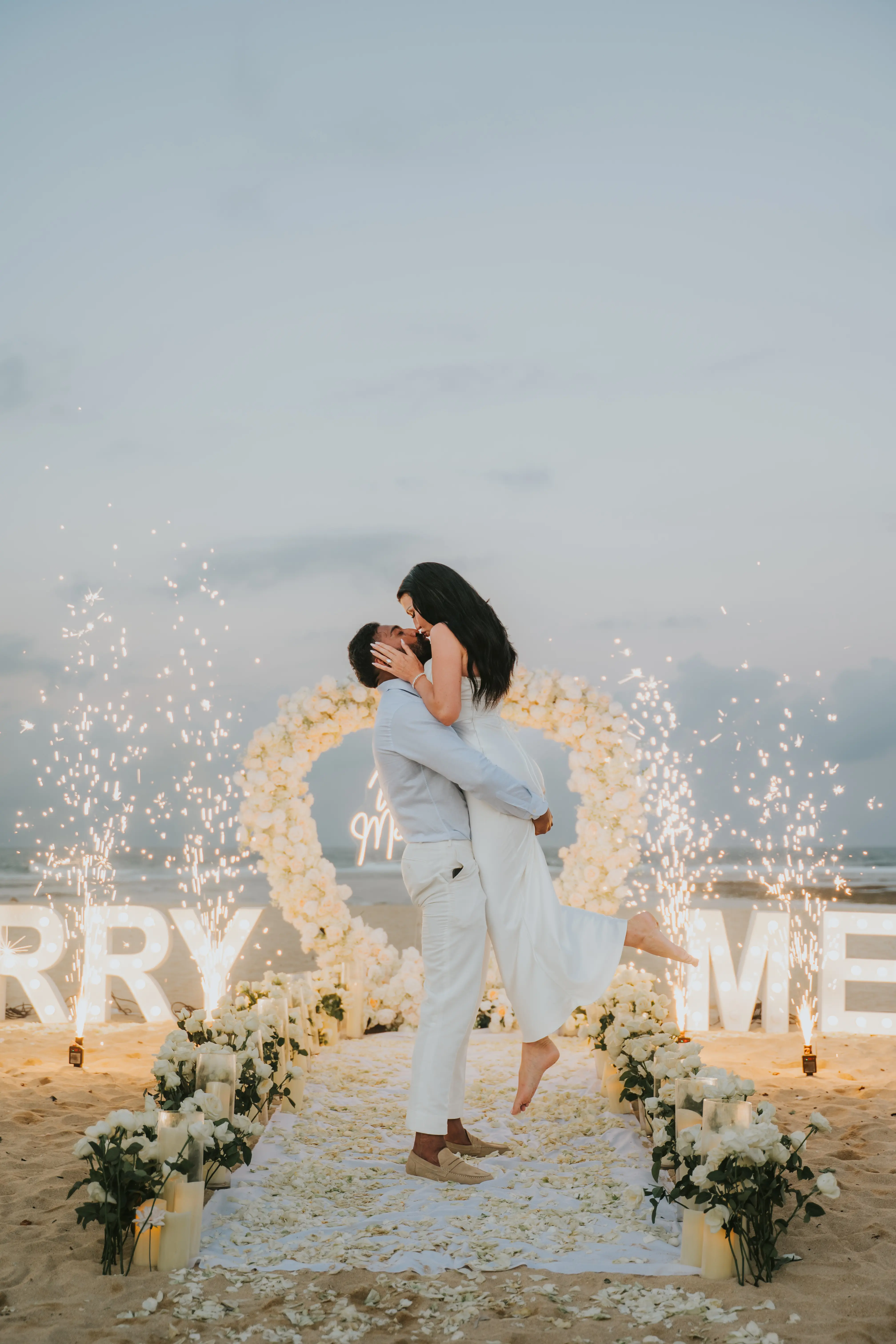 Elegant beach proposal in Bali with white roses and fireworks, groom lifting bride.