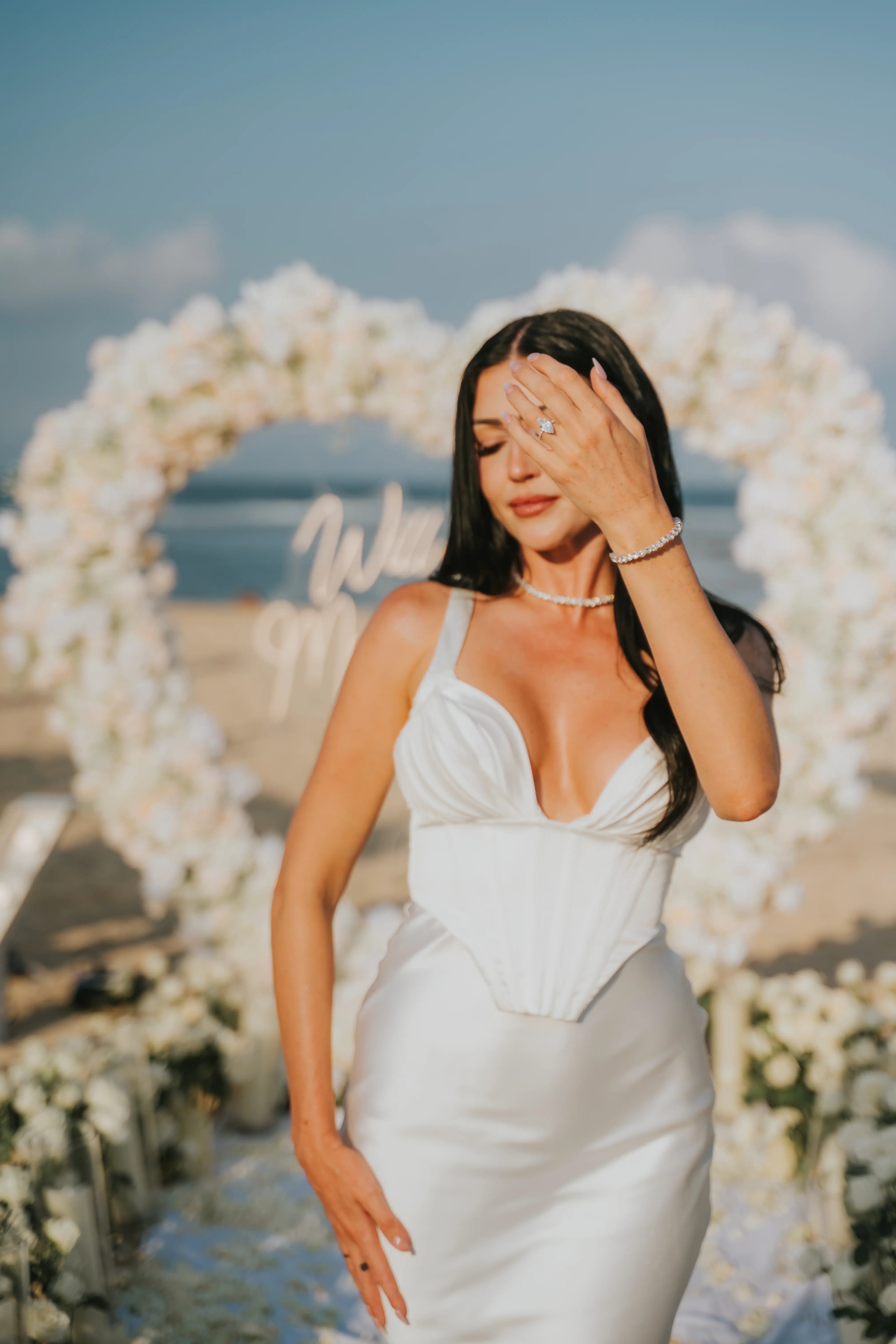 Bride posing in front of white floral heart arch on the beach.