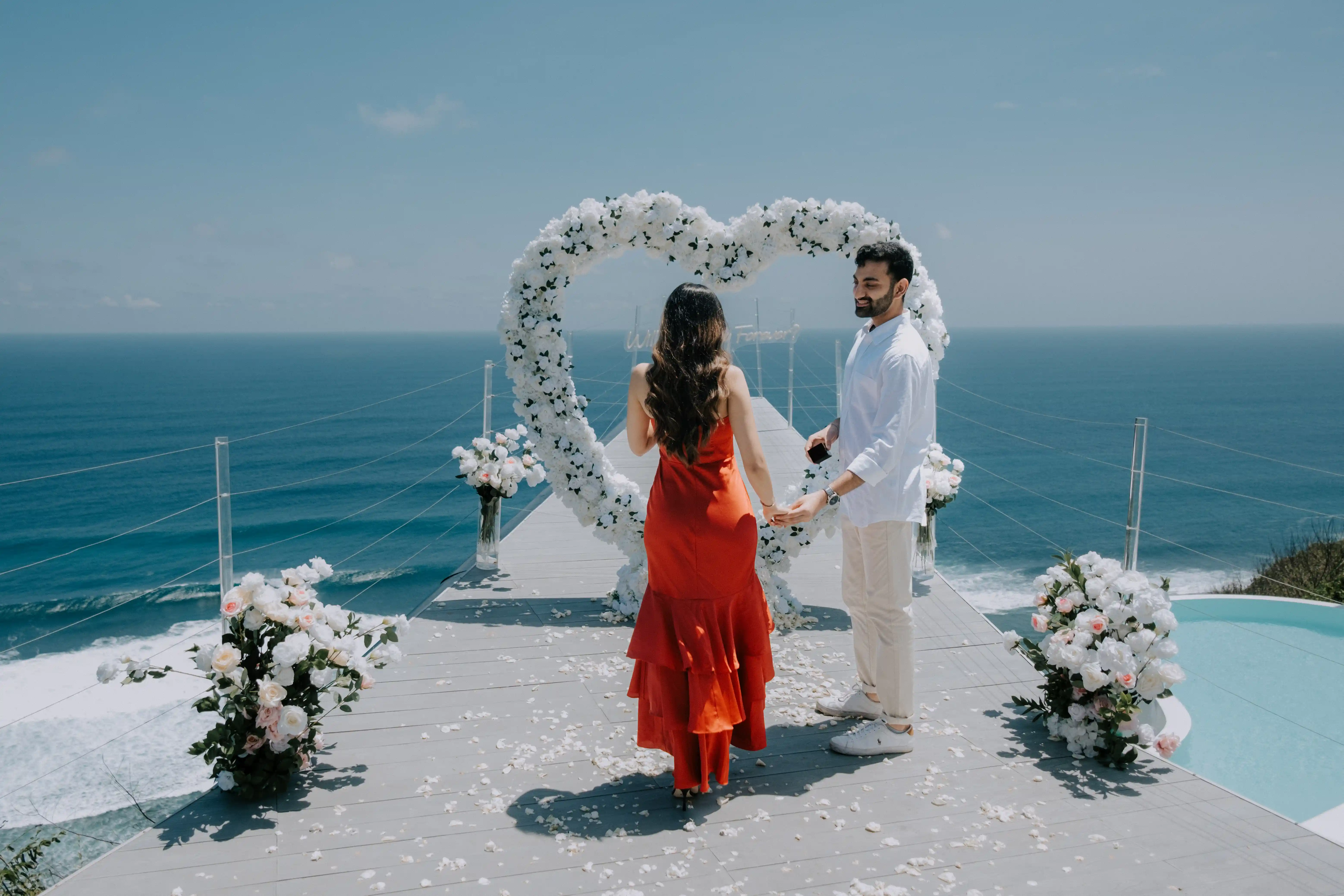 Romantic engagement in Bali with white flower arch and sea backdrop.