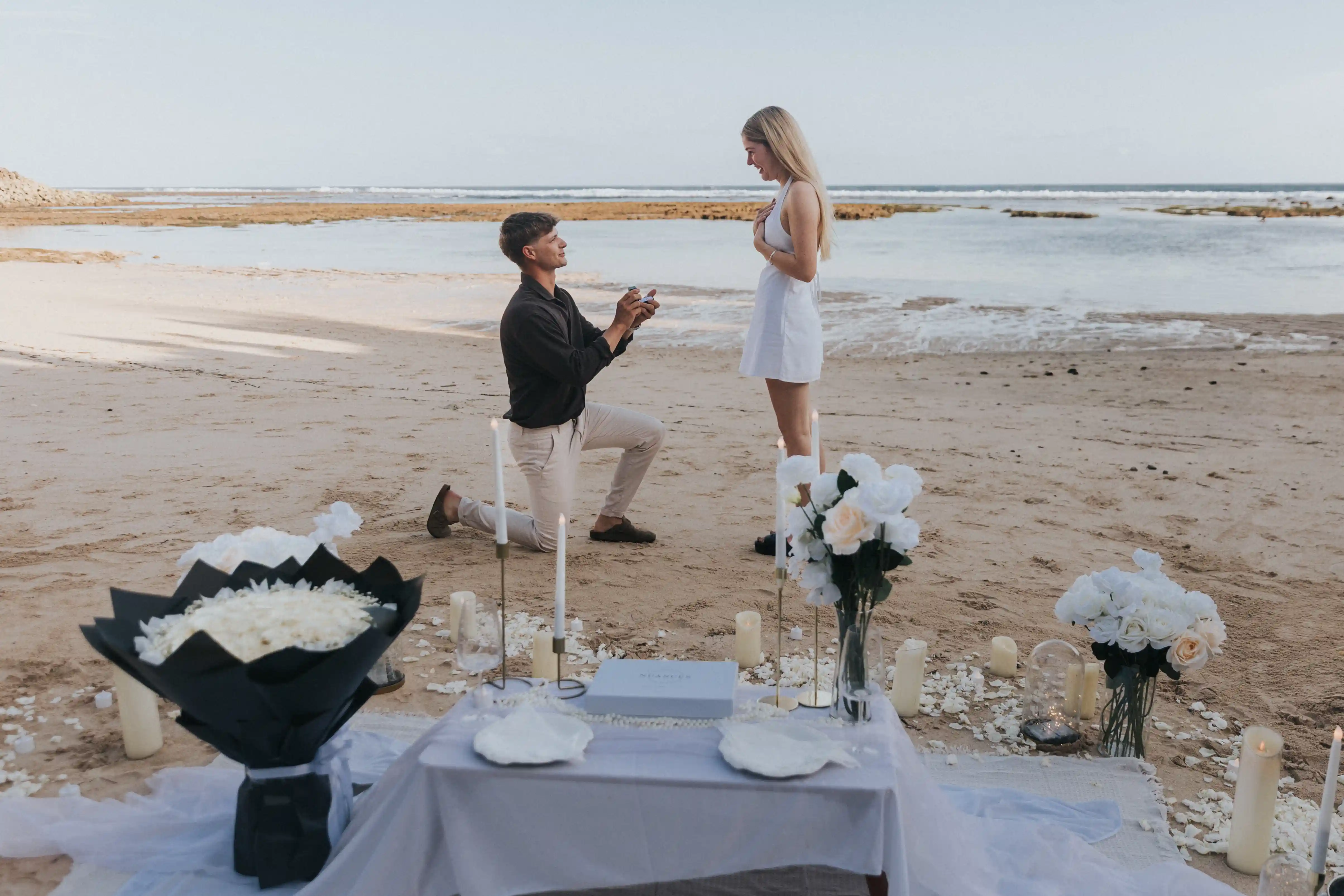 Luxury Bali beach picnic setup with white roses, candles, and ocean breeze — perfect for proposals and romantic dinners.
