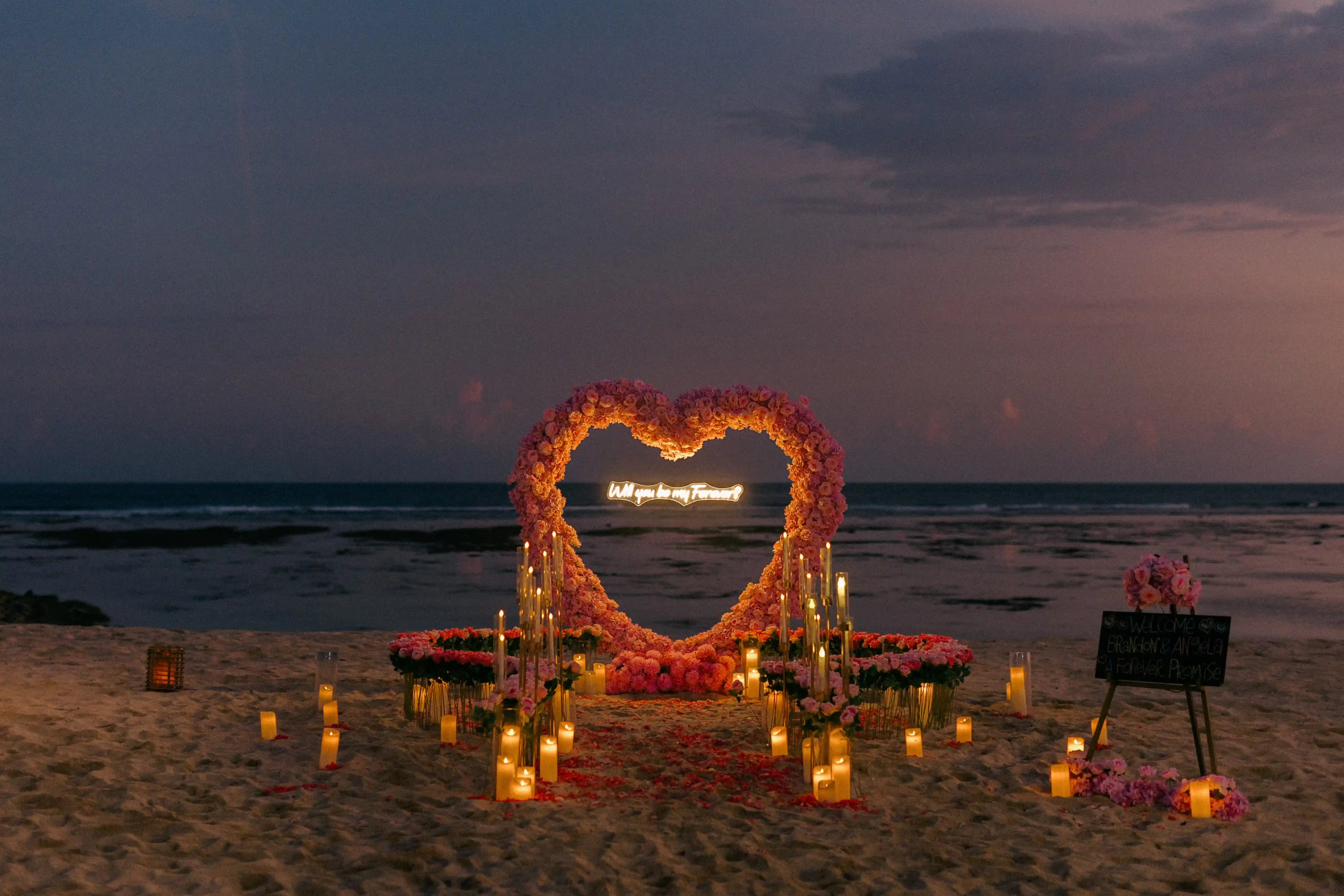 Romantic Bali beach proposal with floral heart arch at dusk.