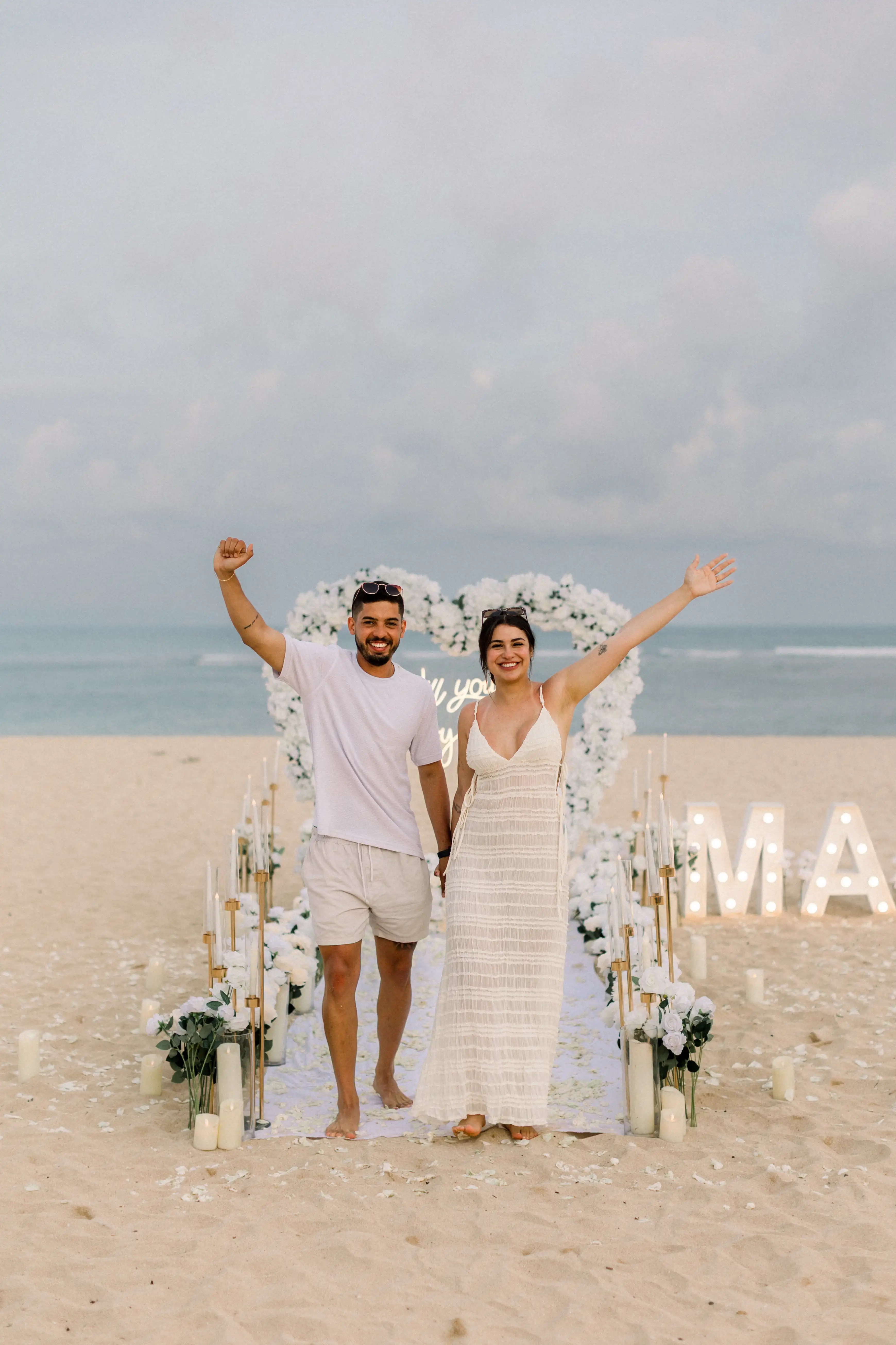 Romantic Bali beach proposal with white heart arch and ocean backdrop.