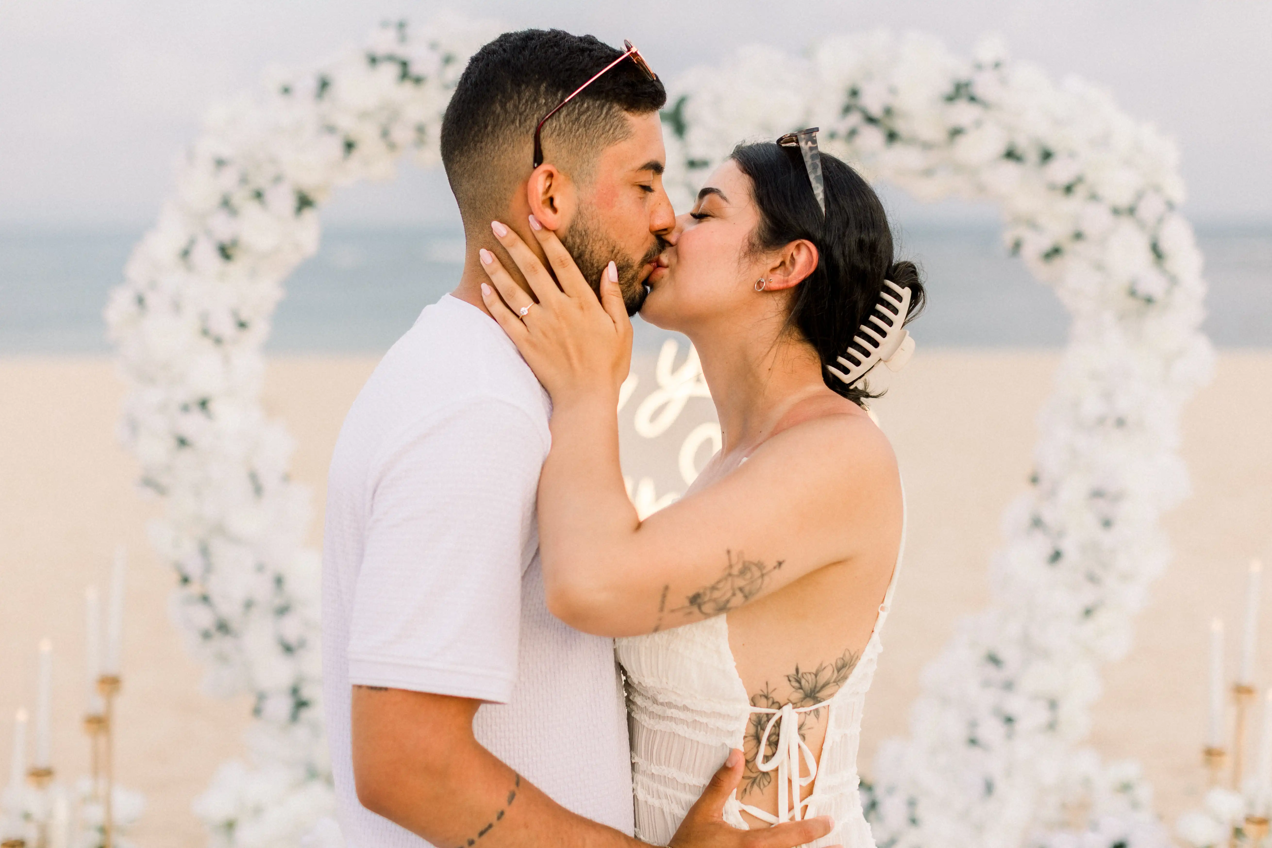 Couple kissing in front of a white heart-shaped flower arch on Bali beach.