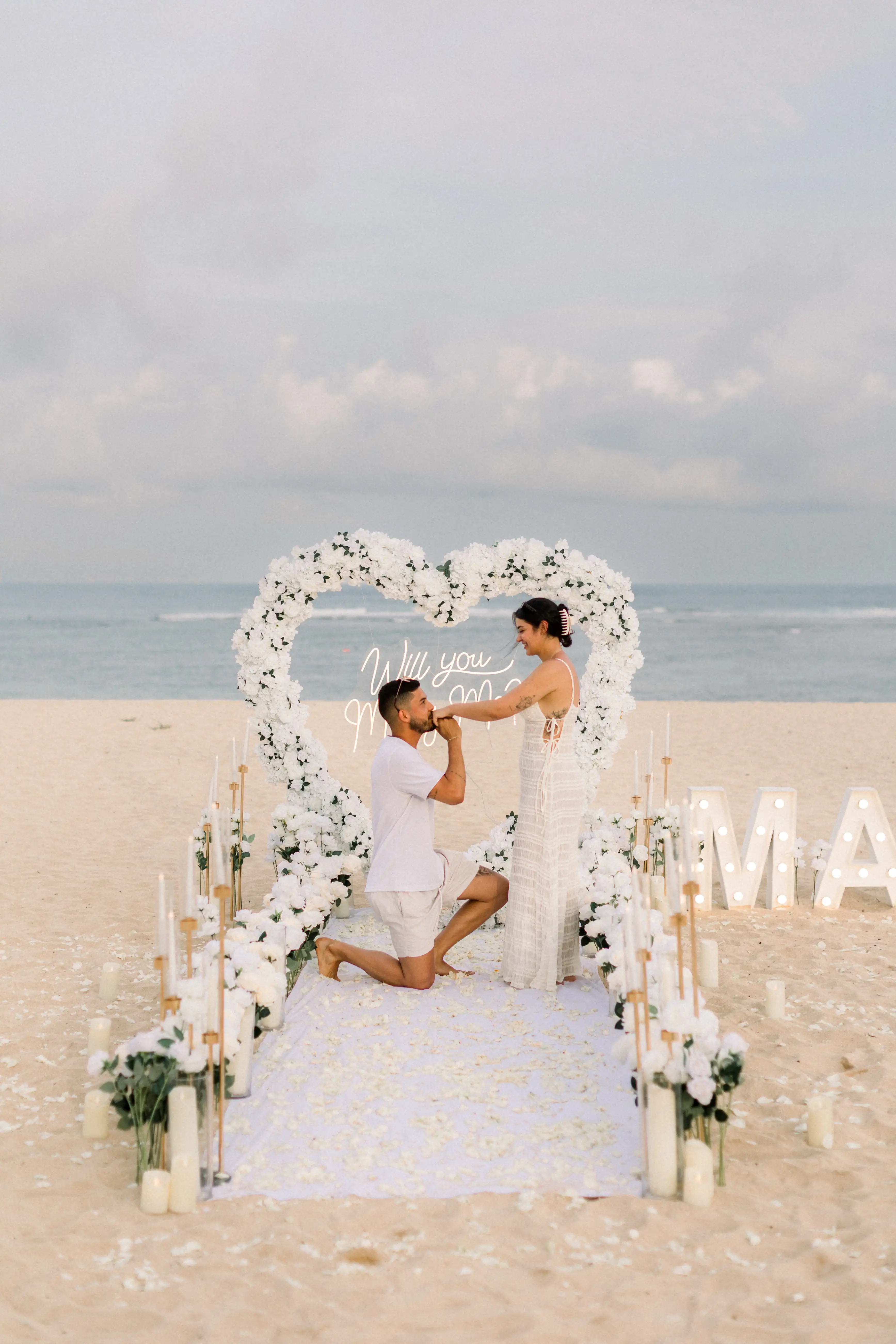 Elegant white floral arch with “Marry Me” letters in the background.