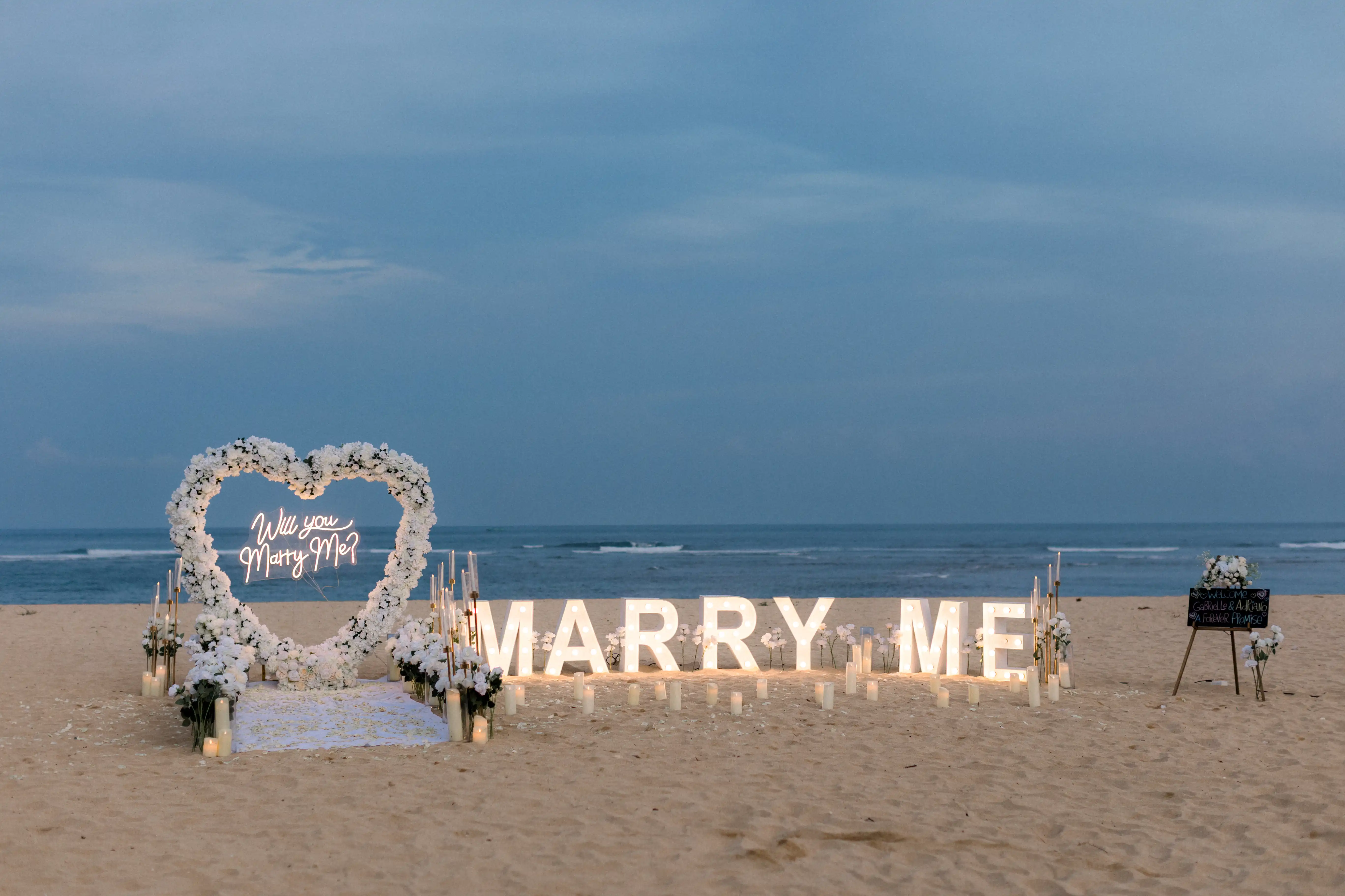 Heart-shaped white floral arch and “Marry Me” sign for a Bali engagement setup.