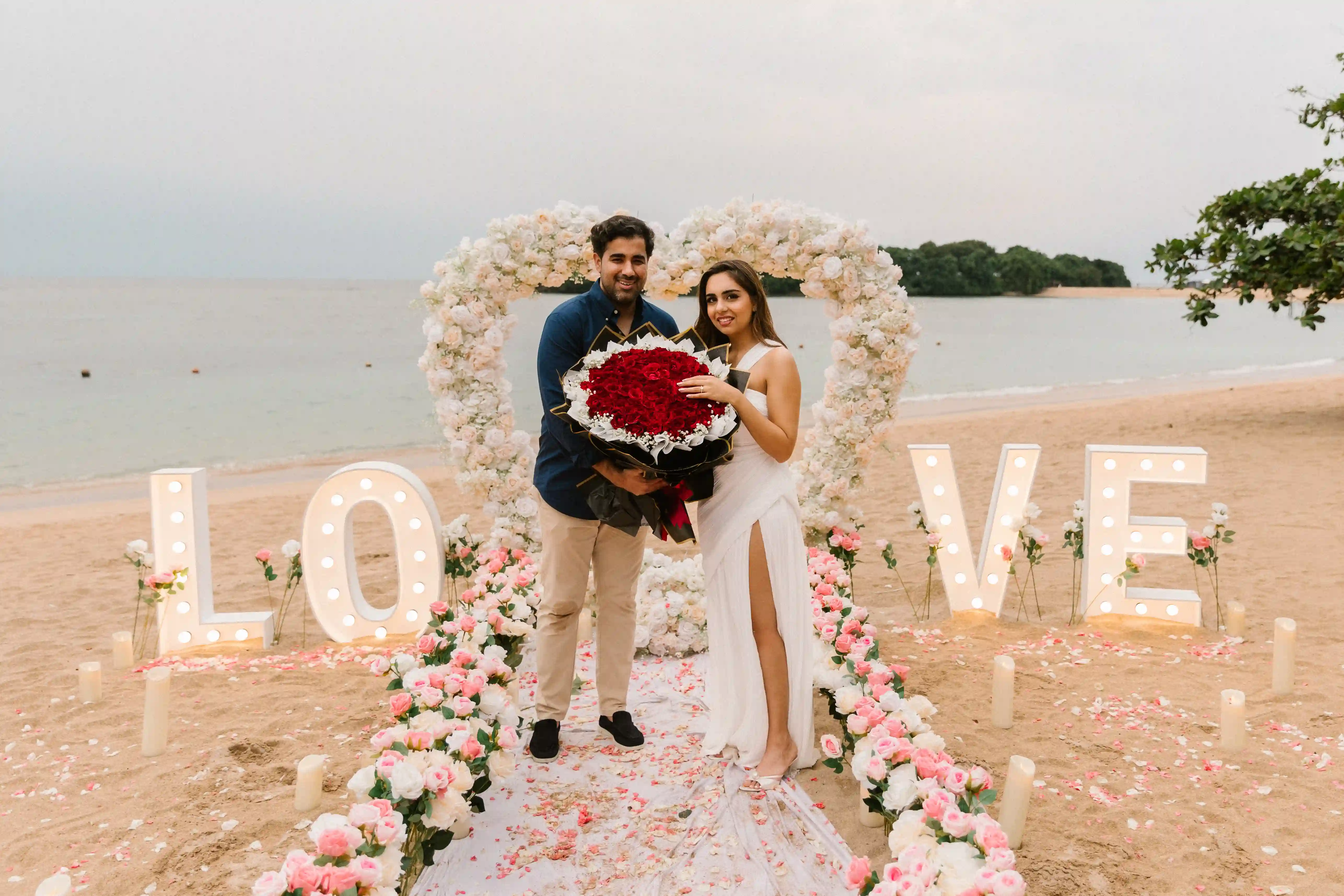 Beach proposal moment framed by floral heart arch and illuminated LOVE letters