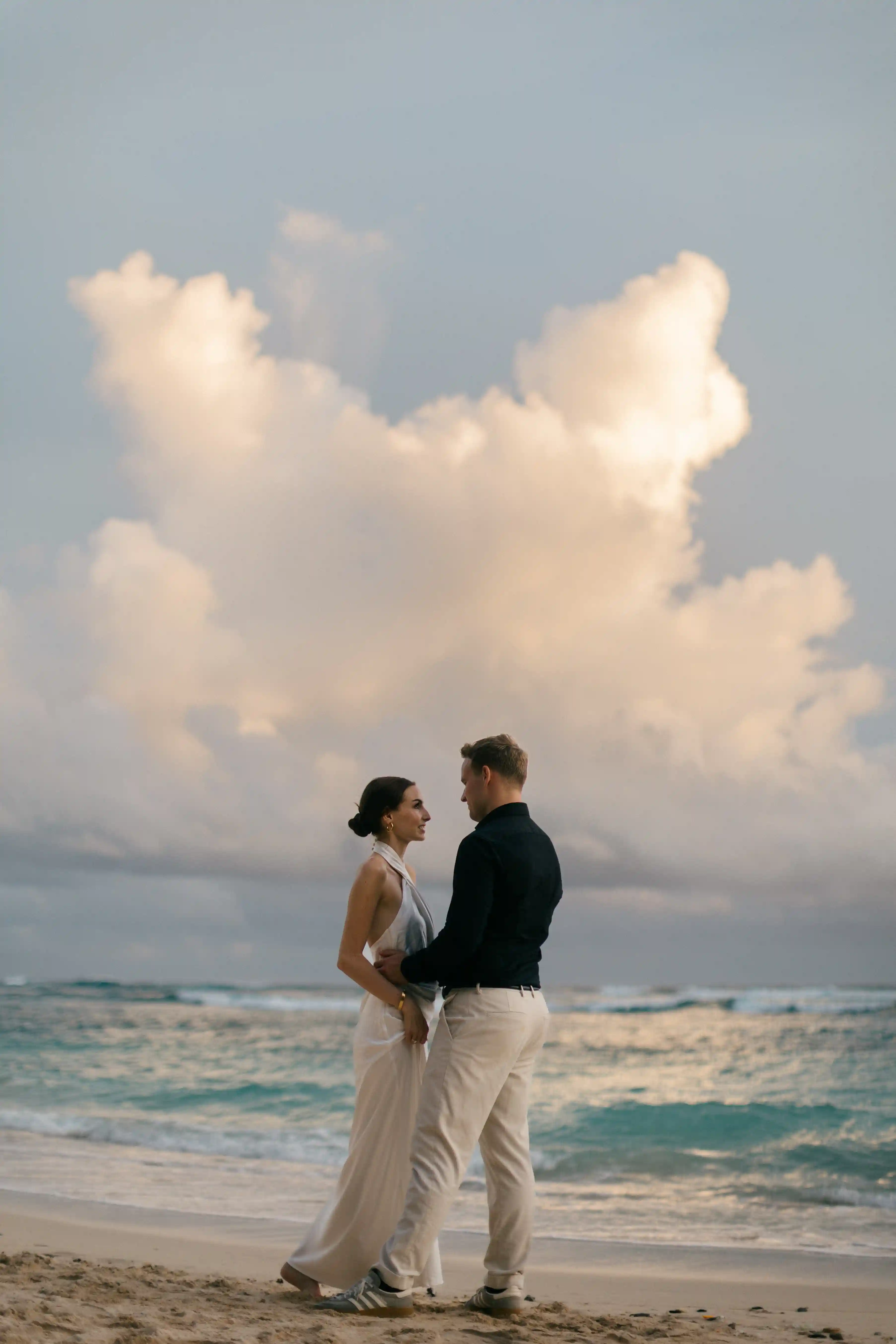 Jasmin and Lucas embracing by the ocean at sunset under a dramatic sky.
