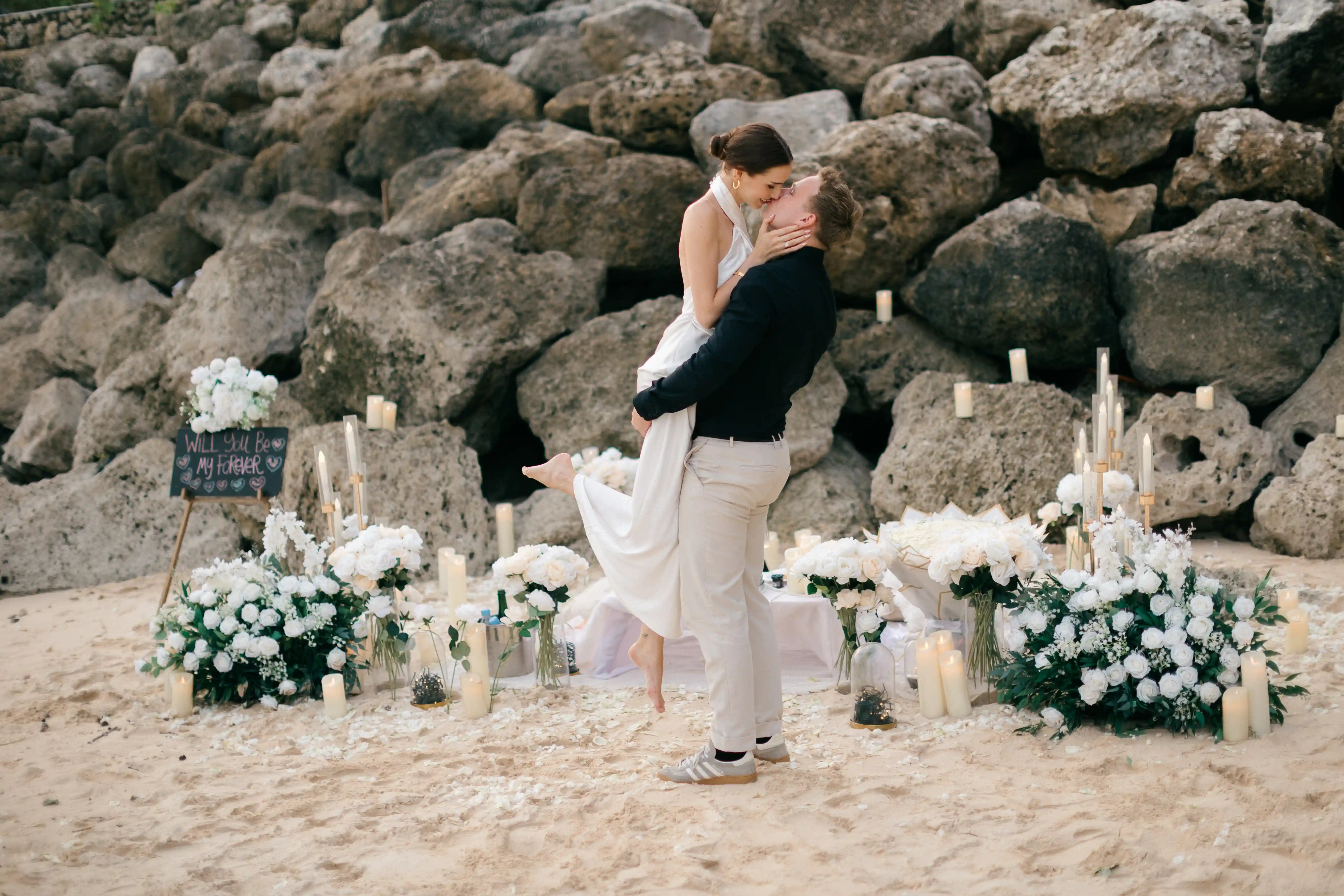 Romantic beach picnic setup with white florals and candles in front of natural rocks.