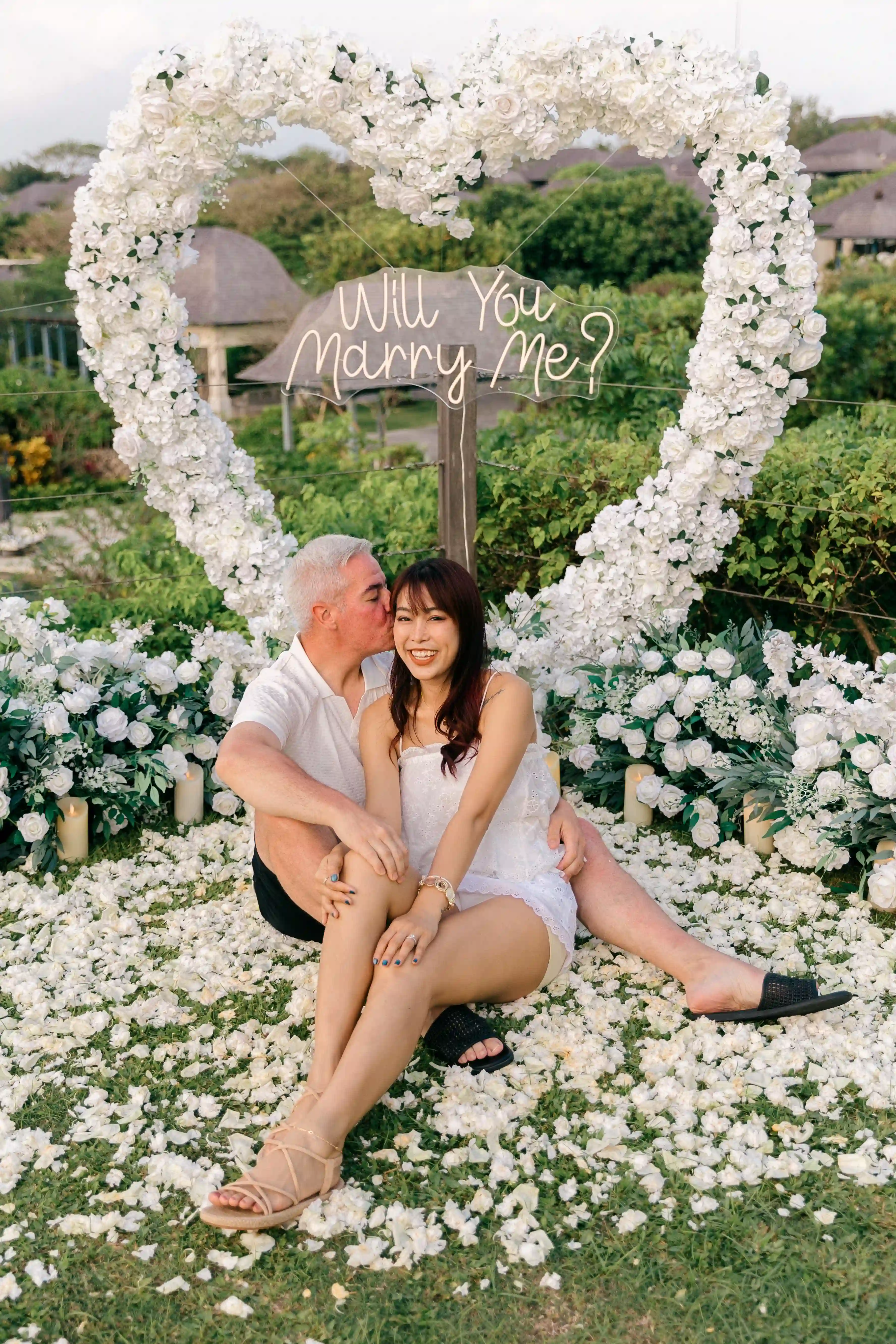 Happy couple sitting in front of floral heart arch after proposal in Bali