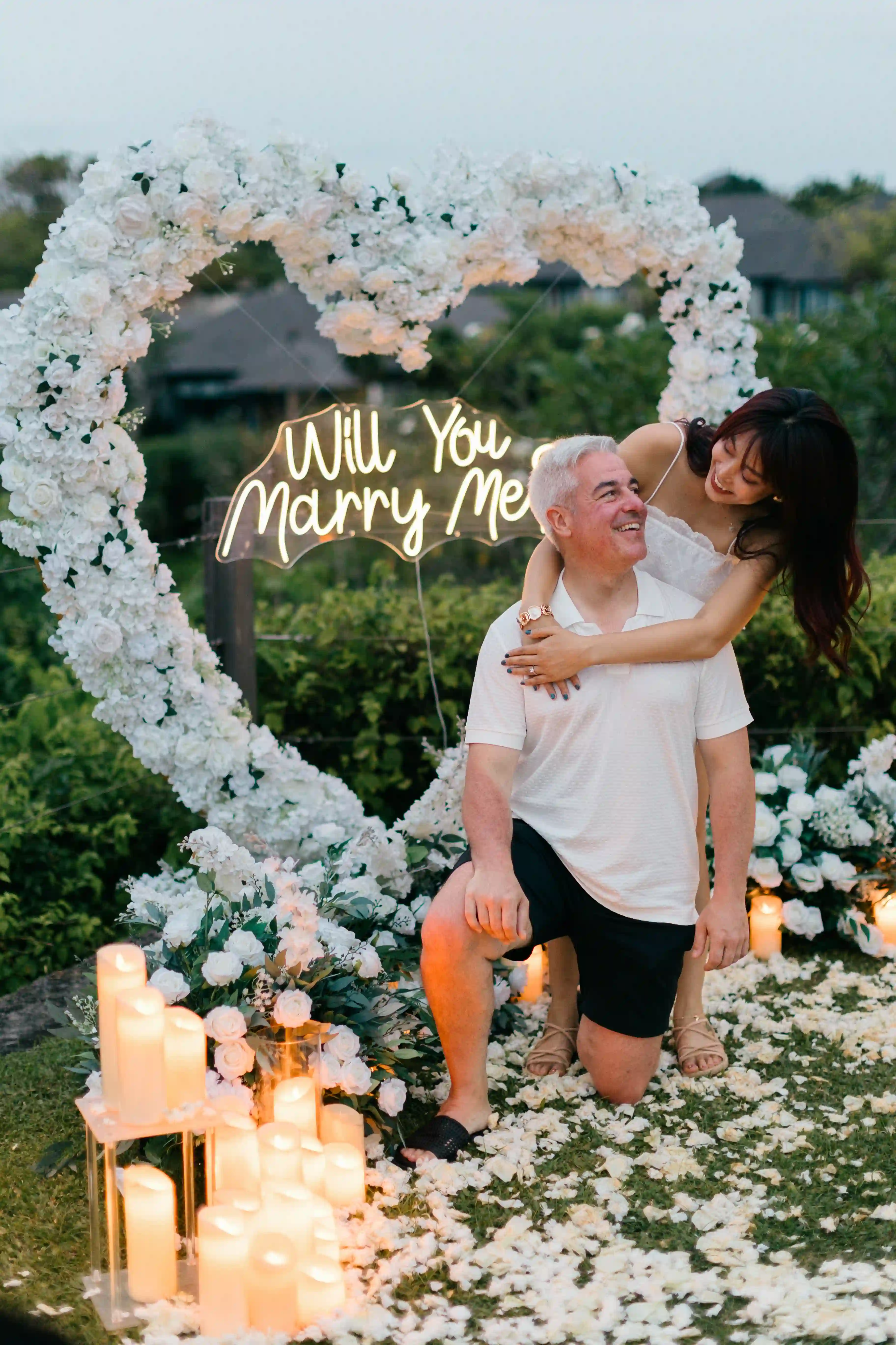 Couple celebrating engagement in front of white floral heart arch in Bali