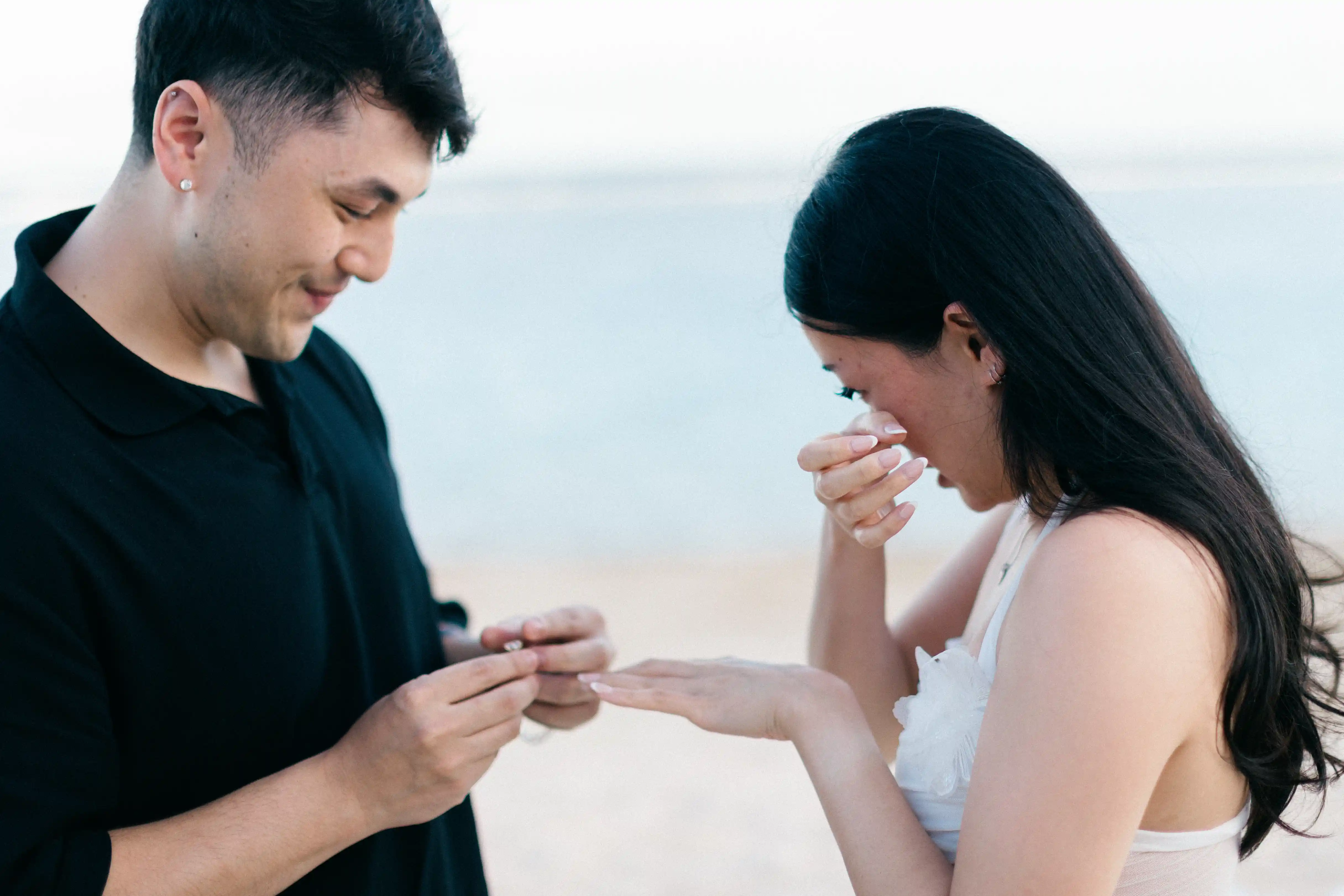 Groom placing engagement ring on bride’s finger during beach proposal