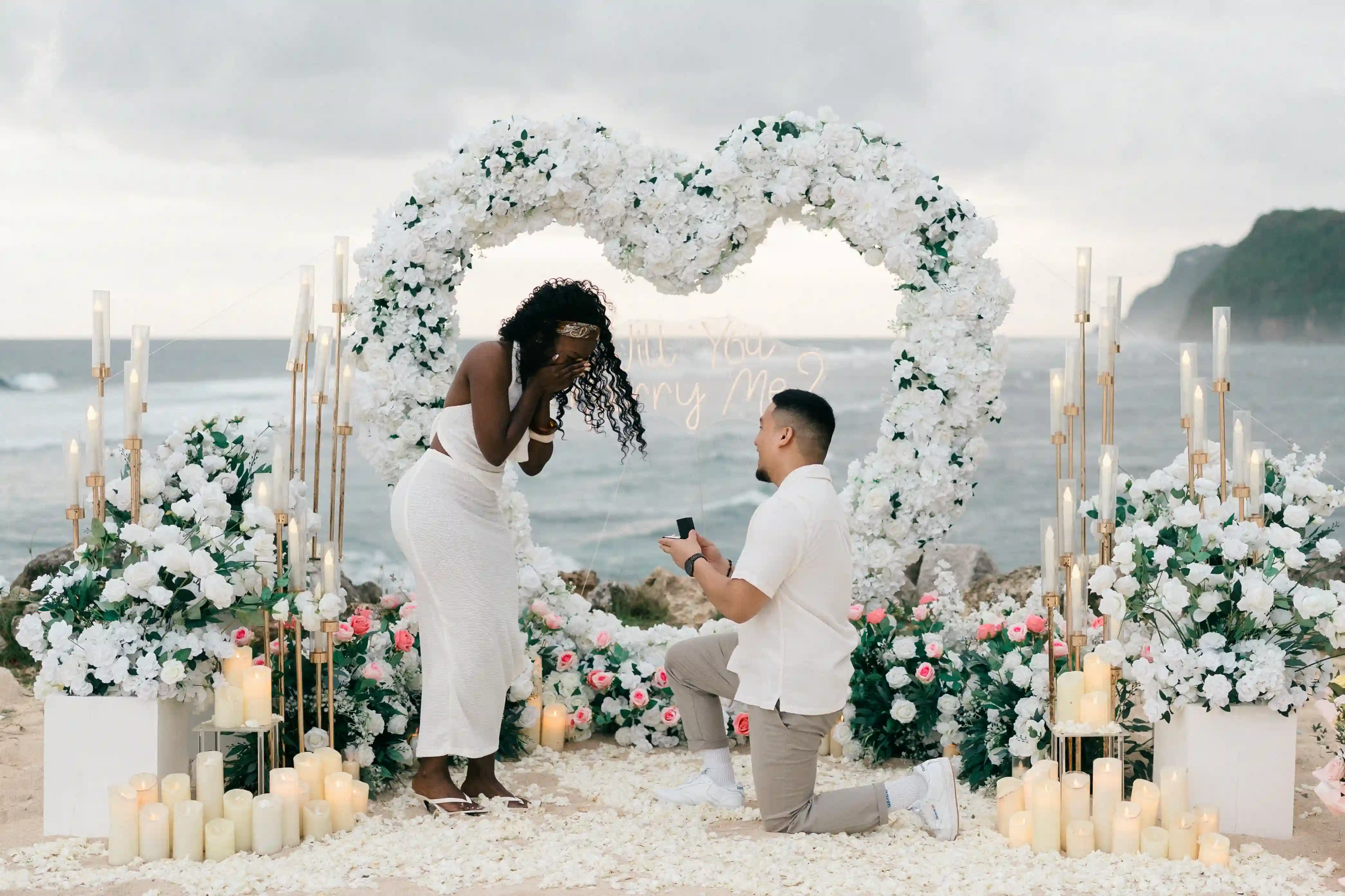 Man proposing beneath white floral heart arch during Bali beach proposal