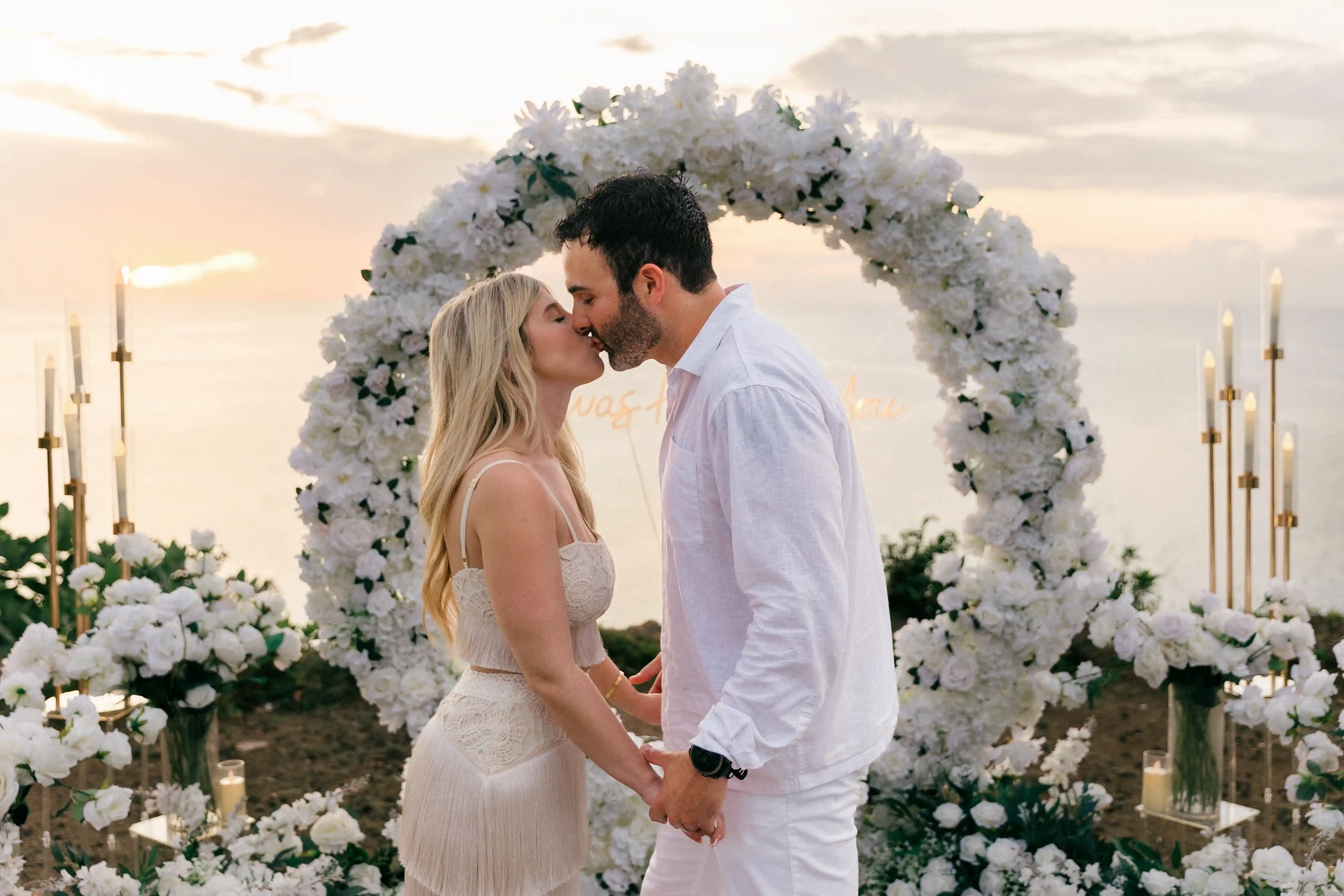 Couple standing beneath white floral arch during cliffside Bali proposal