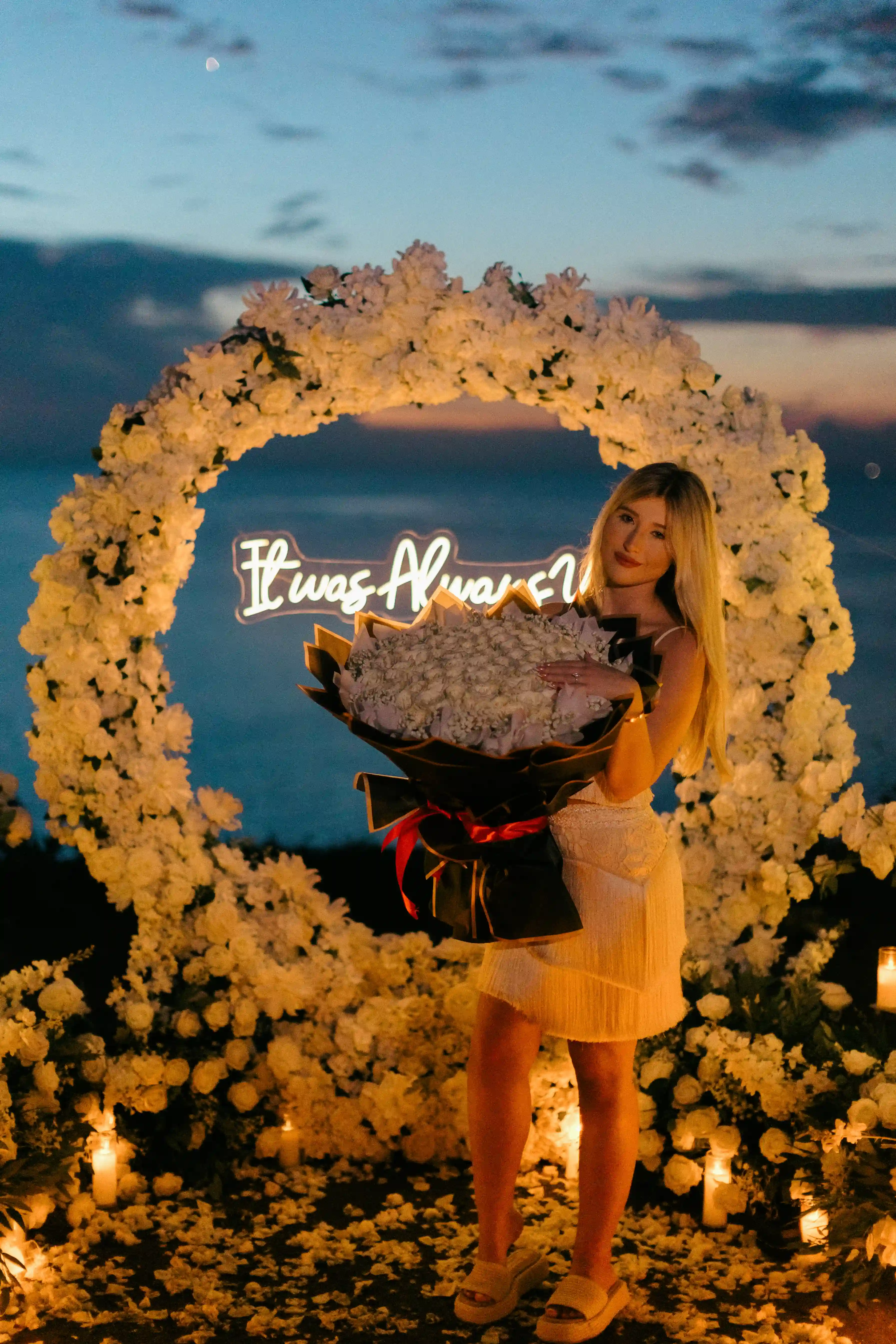 Bride showing engagement ring in front of floral arch and neon sign