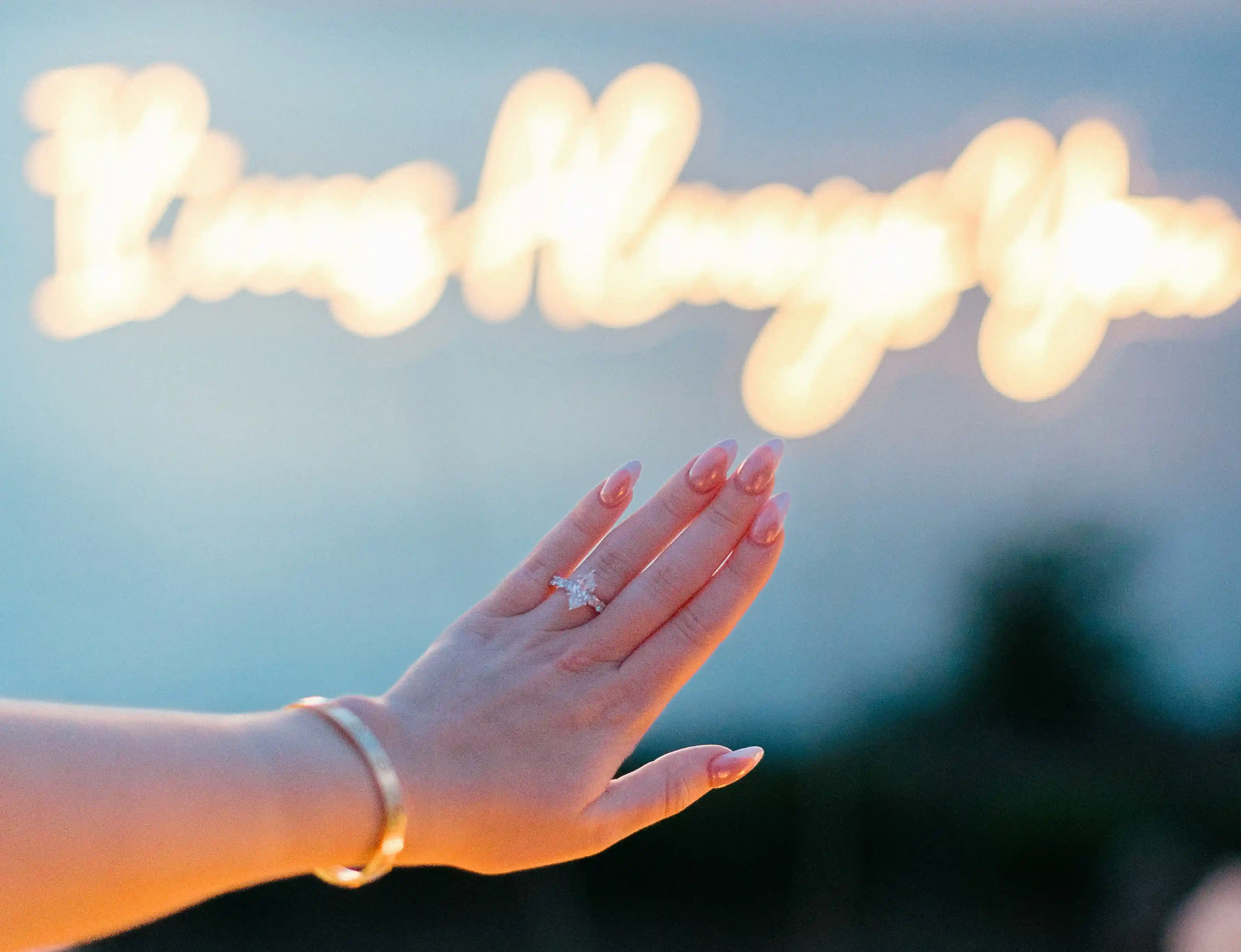 Close-up of engagement ring in front of blurred “It was Always You” neon sign