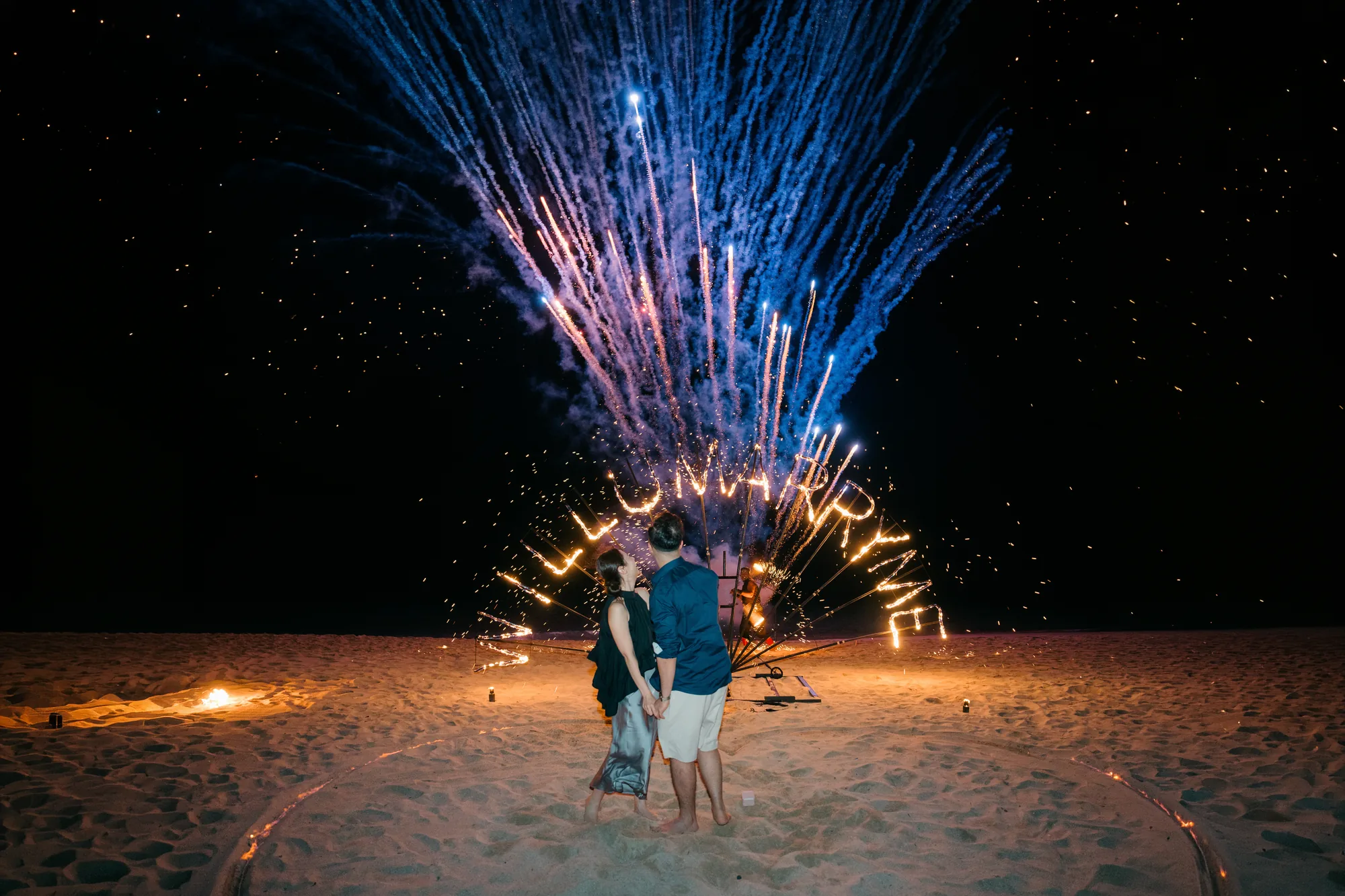 Couple at a beach proposal in Bali with blue fireworks bursting overhead
