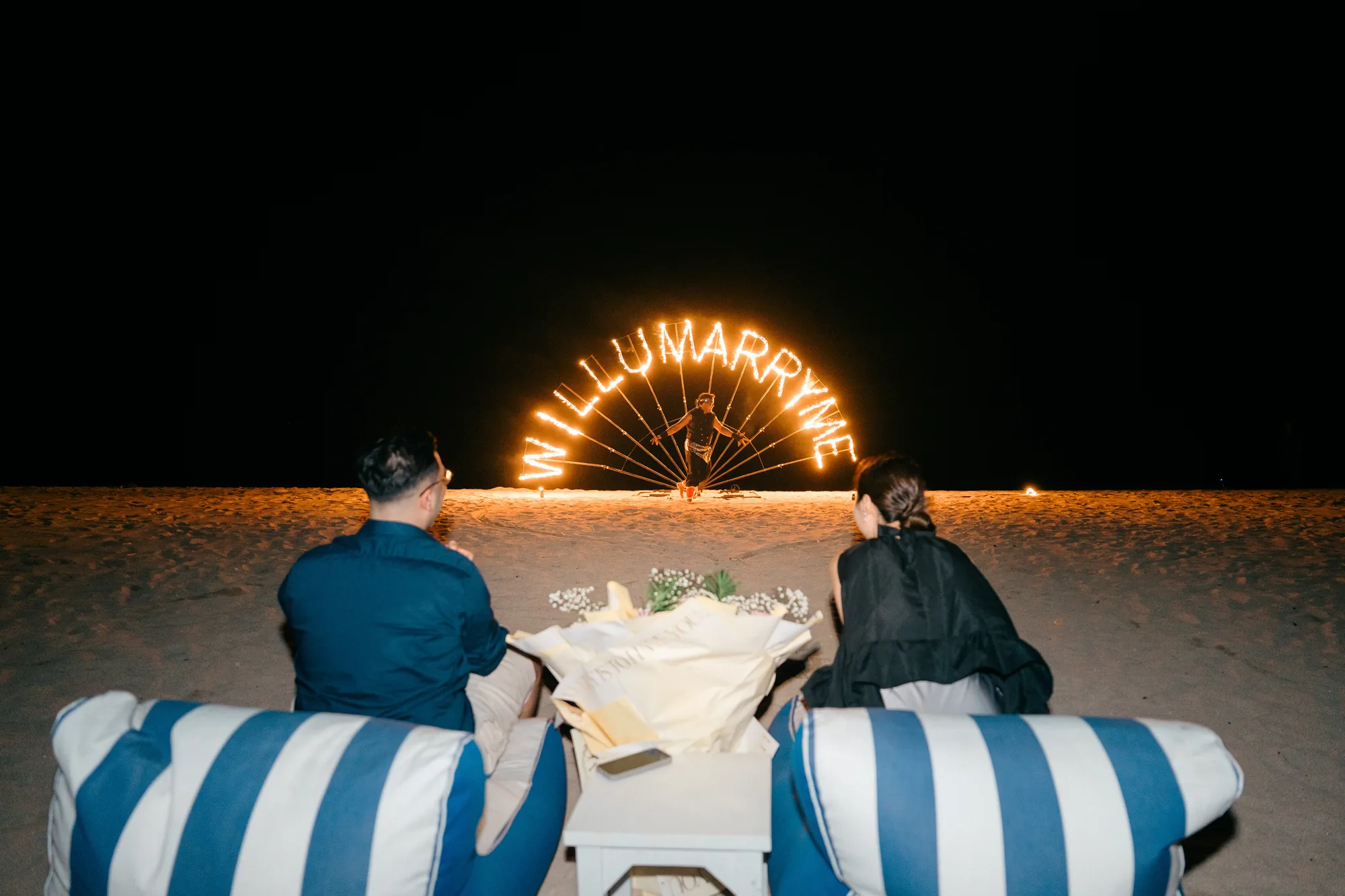 Couple watching the 'Will U Marry Me' fire letters being lit at a Bali beach proposal