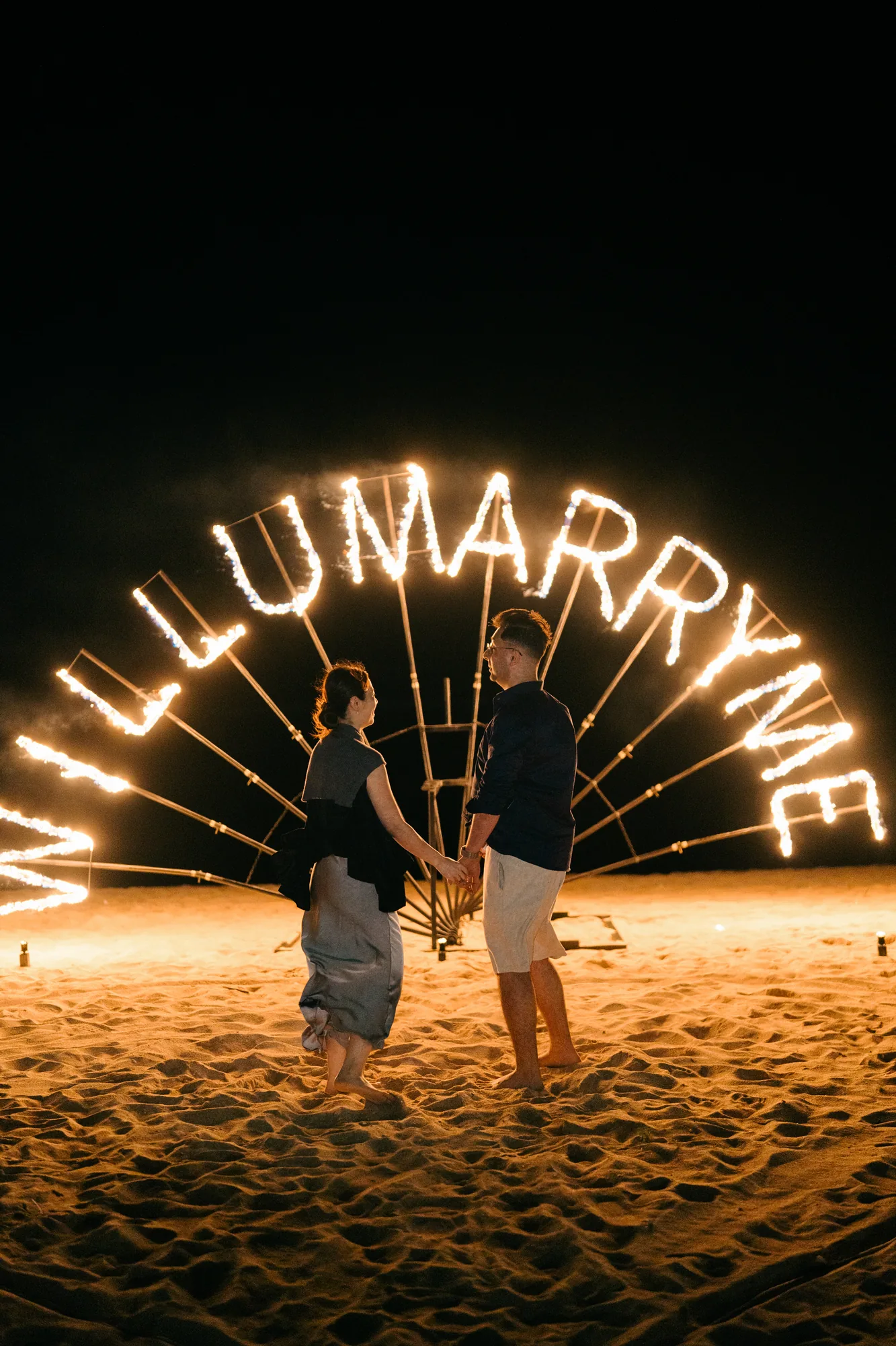 Couple holding hands in front of a 'Will U Marry Me' fire sign at a Bali beach proposal