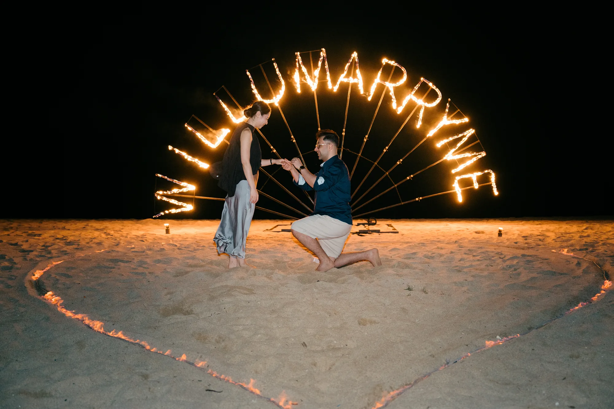 Man kneeling to propose in front of 'Will U Marry Me' spelled in fire on the beach in Bali