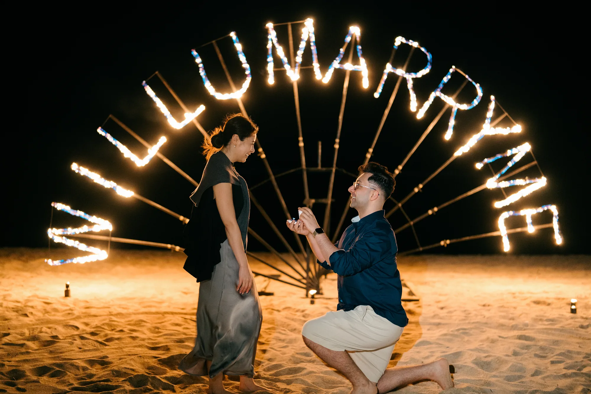 Couple at the moment of proposal by the 'Will U Marry Me' fire sign on a Bali beach