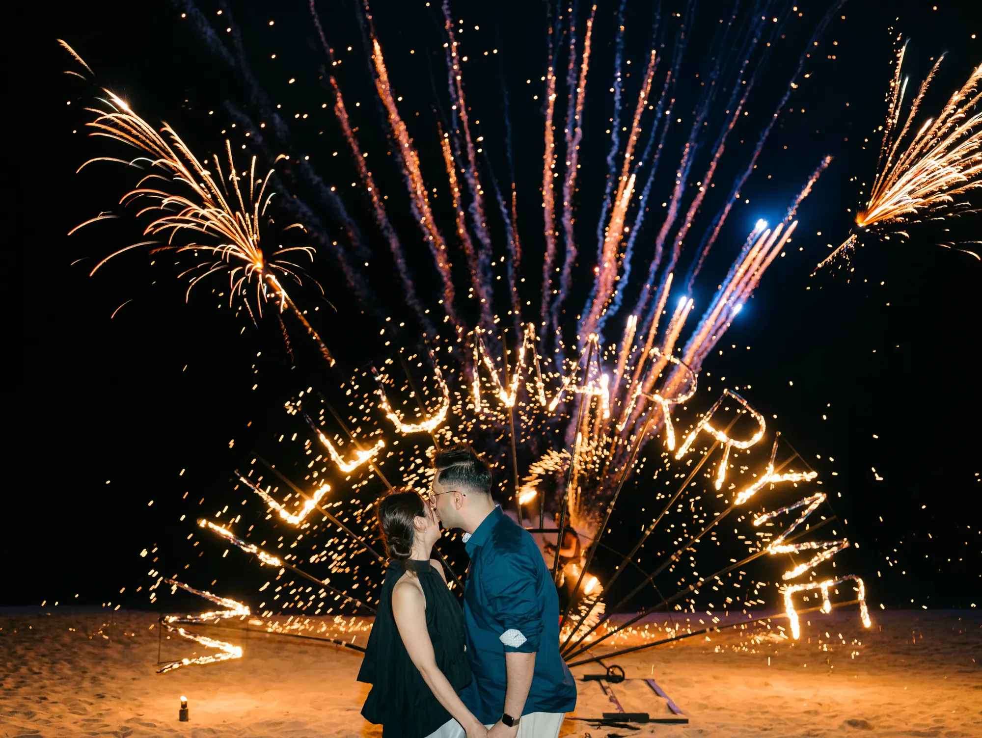 Couple embracing as fireworks light up a 'Will U Marry Me' fire sign on a Bali beach