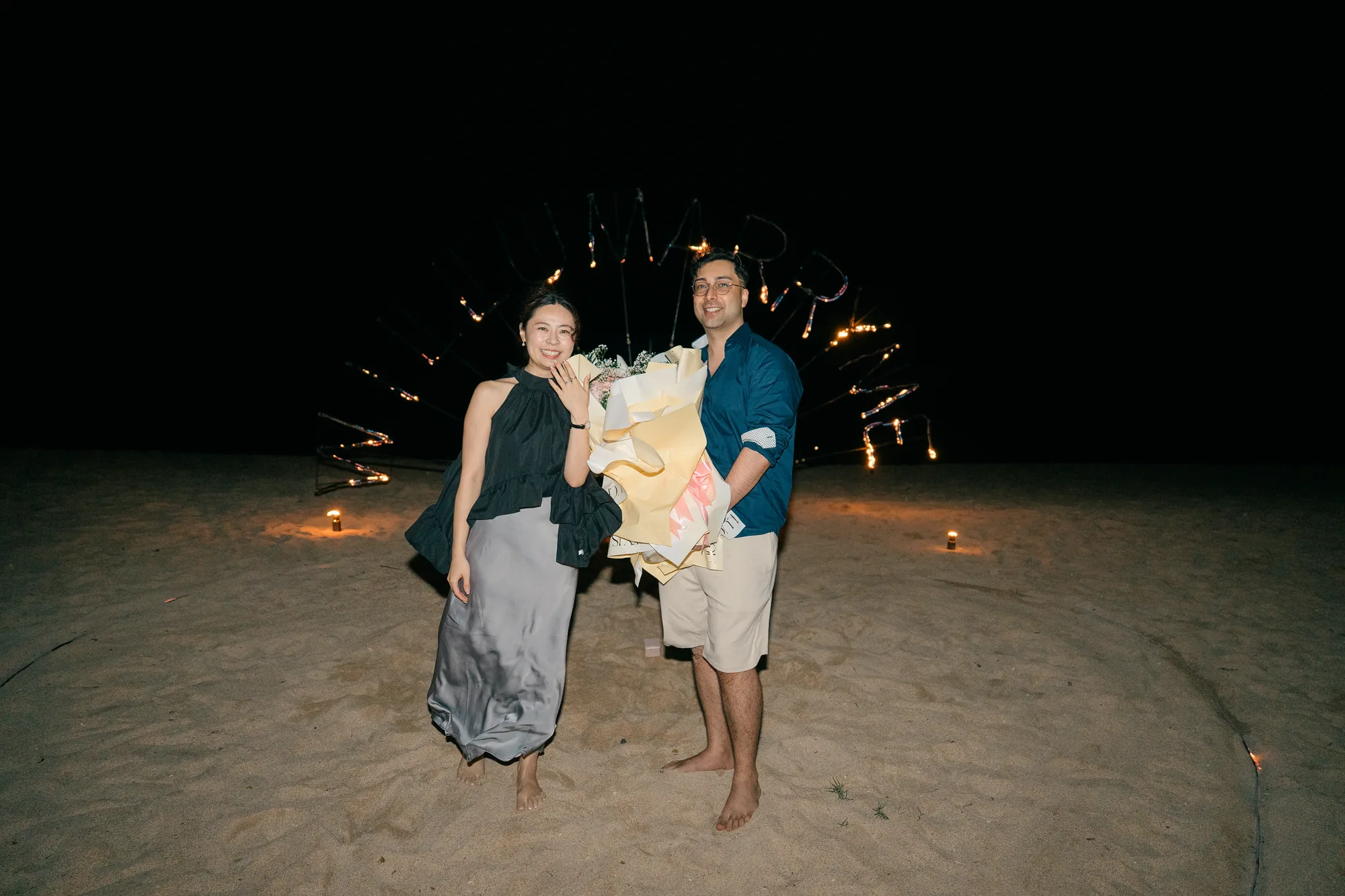 Couple on the beach holding a bouquet with sparklers glowing behind at a Bali proposal